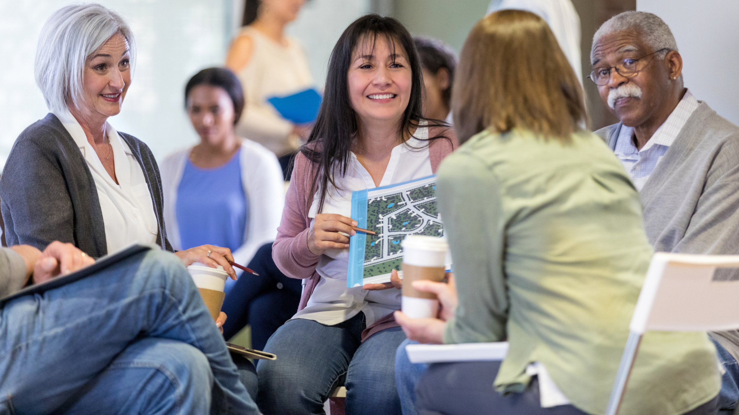 A group of adults sits in a circle having a discussion about human services networking opportunities; one woman holds a map or site plan, and others hold coffee cups.
