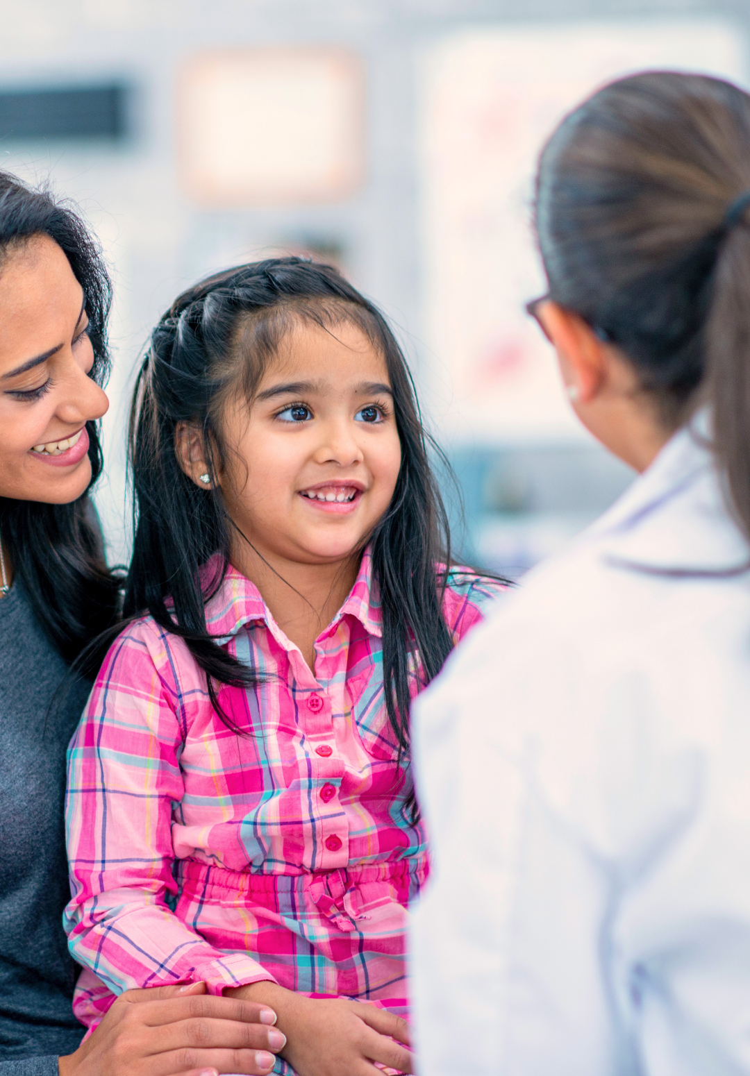 A young girl with long dark hair and a pink plaid shirt is smiling while talking to a woman in a white lab coat, with another woman sitting beside her holding her hand in a medical or healthcare setting.