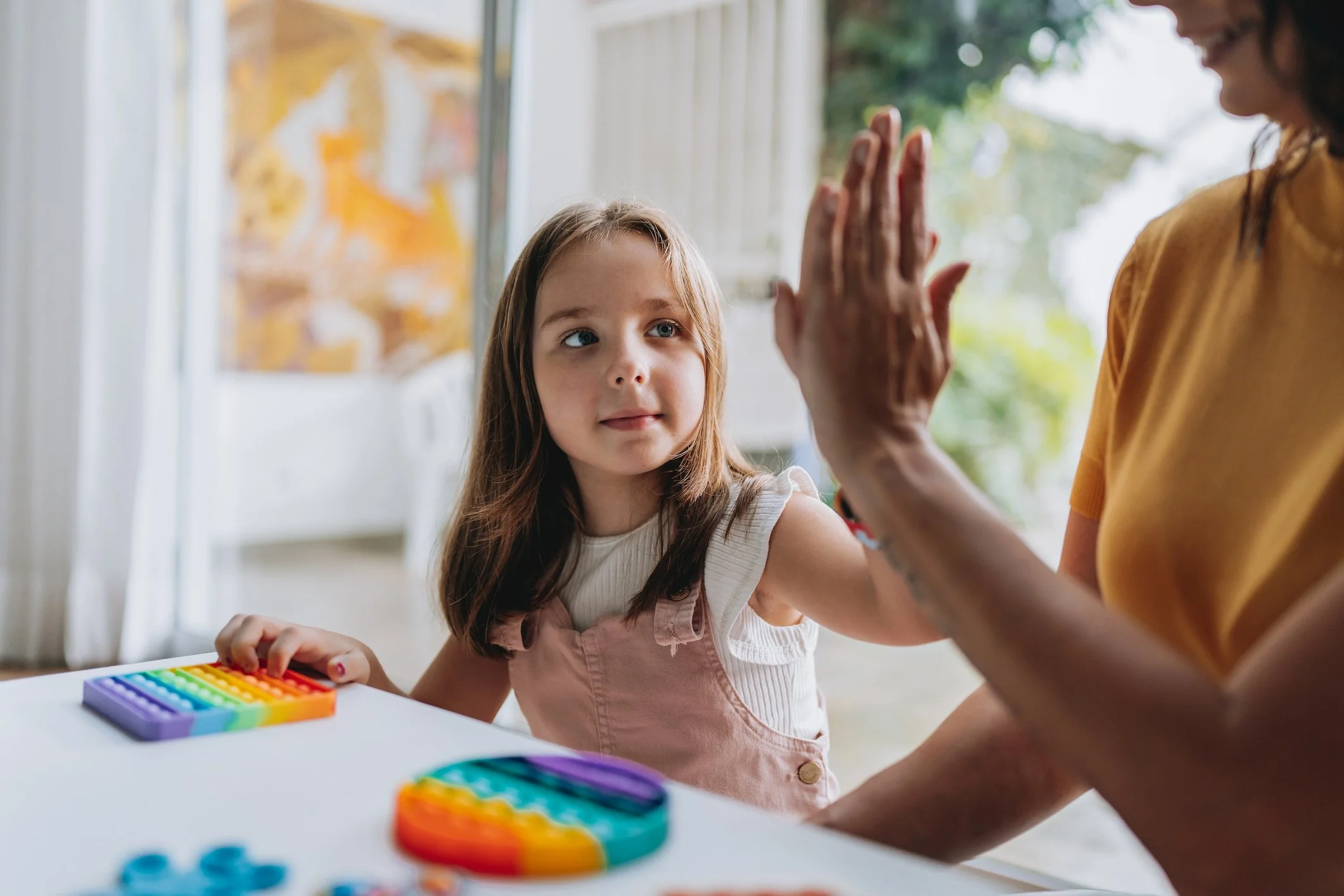 A young girl with brown hair and a white shirt sits at a table with colorful sensory toys, while an adult high-fives her. The background shows a window with outdoor scenery.