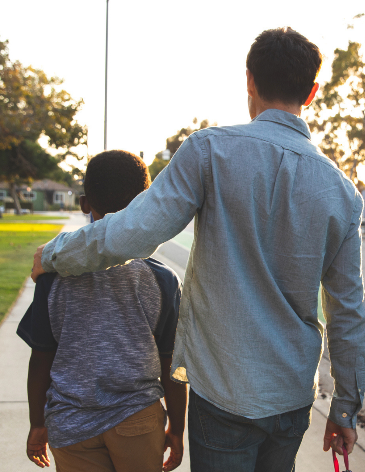 An adult with their arm around a child walks down a sidewalk in a suburban neighborhood during the day showing foster care best practices.