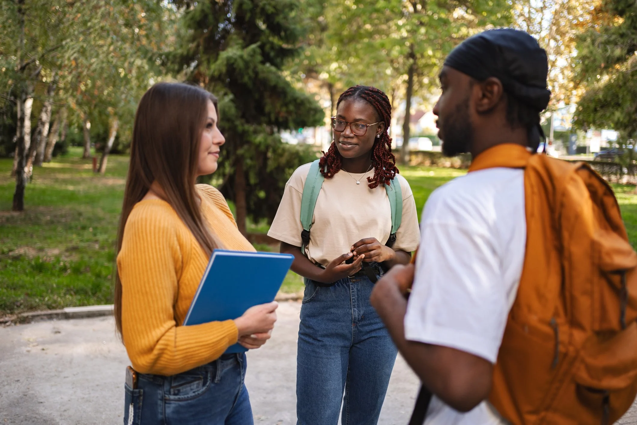 Three young adults standing outdoors in a park, engaged in conversation. The woman on the left holds a blue notebook, the woman in the center wears glasses and a backpack, and the man on the right has a backpack and a bandana.