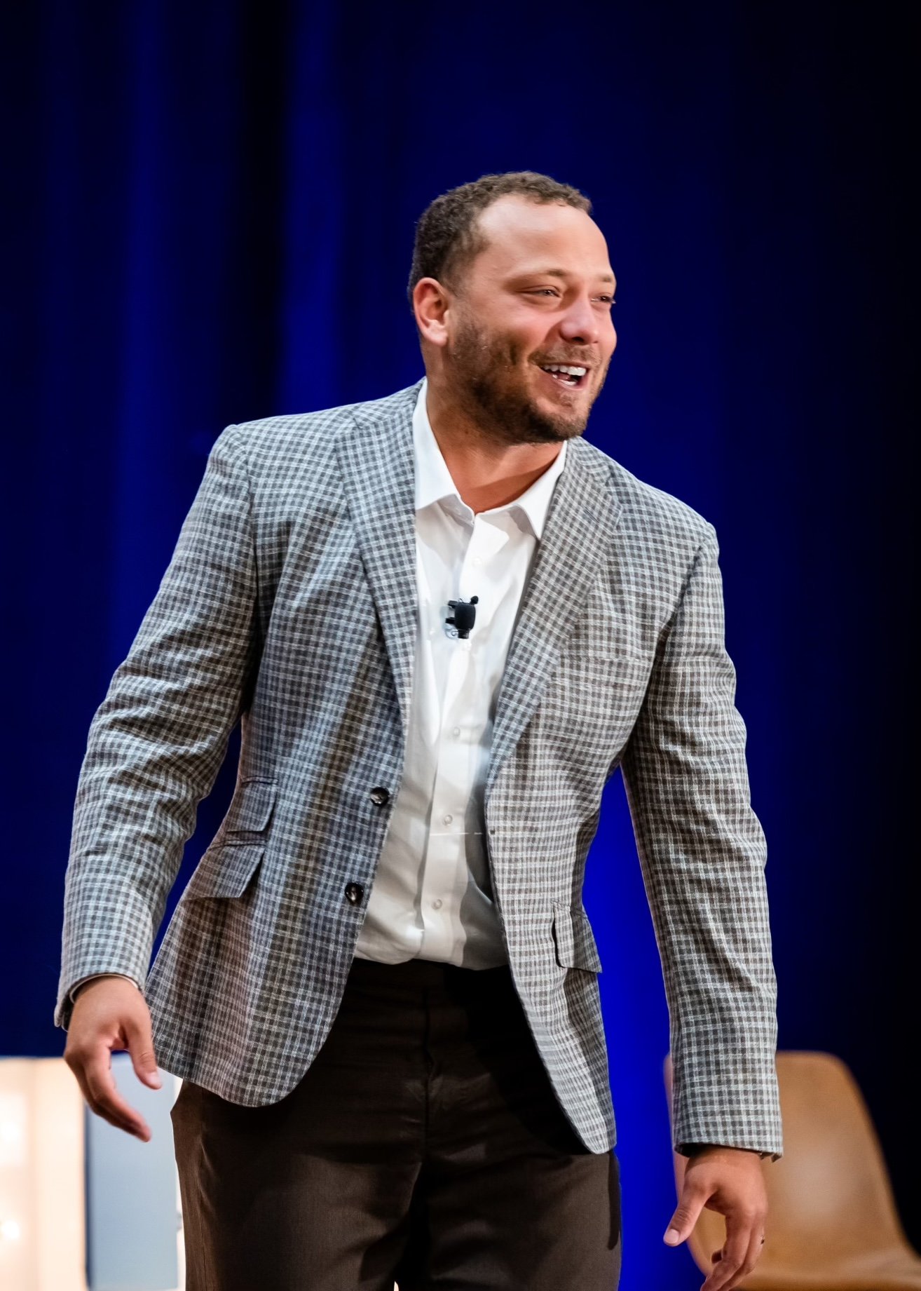 Alec Ingold, a FFTA guest speaker in a checked blazer and white shirt stands and smiles on stage with a blue curtain backdrop.