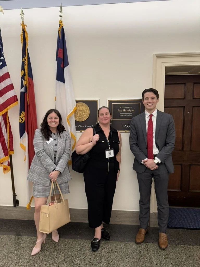 Three child welfare advocates pose in business attire in front of an office door and several flags inside a government building.