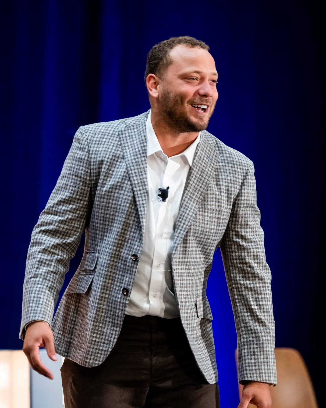 Alec Ingold in a gray checkered blazer and white shirt speaks on stage with a microphone, smiling against a blue background.