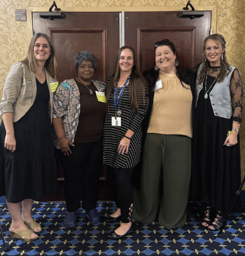 Five women stand together indoors, smiling at the camera in front of double wooden doors for family support programs