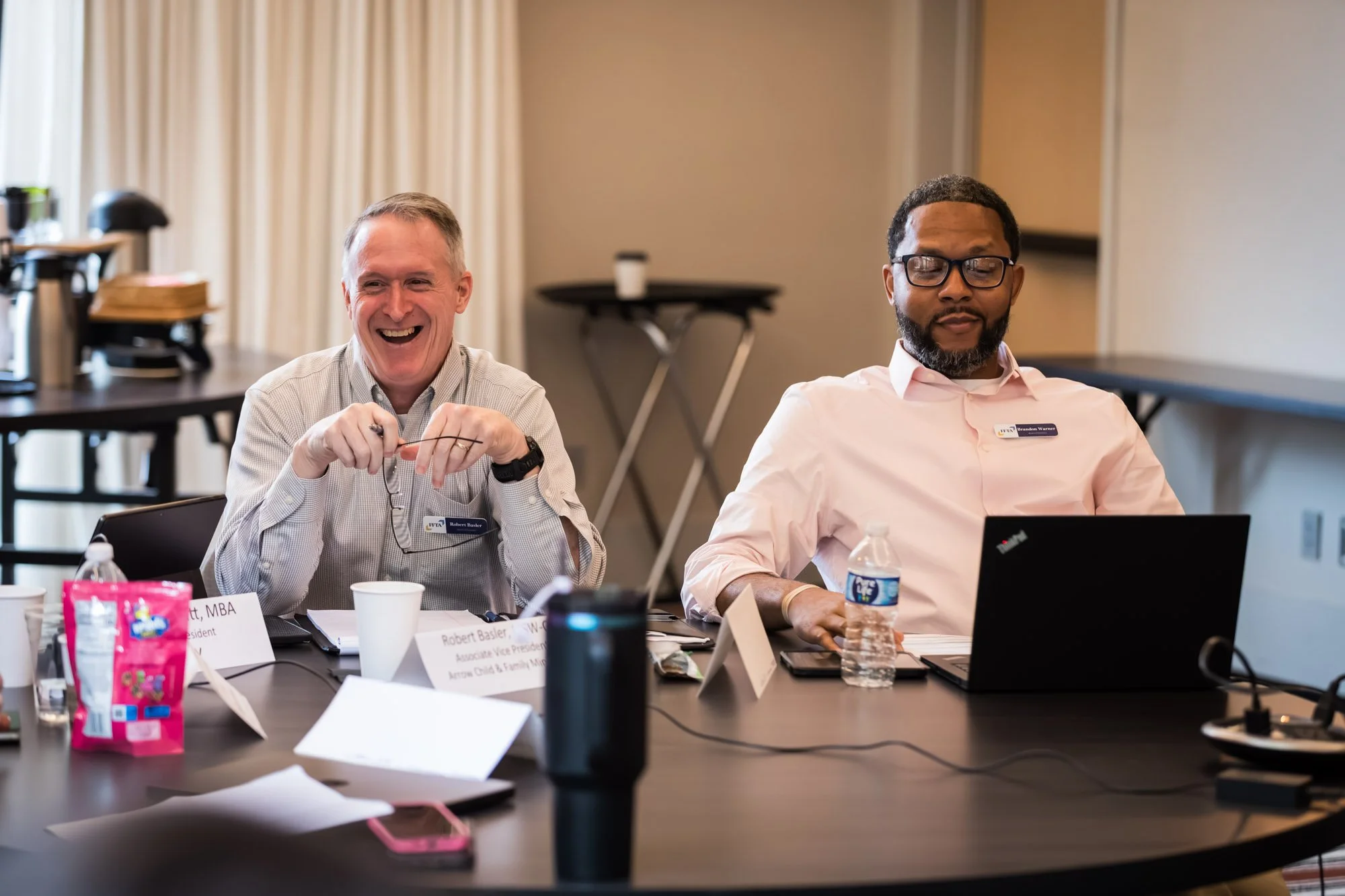 Two men sit at a conference table with laptops and papers, one smiling, during a meeting about family-serving organizations