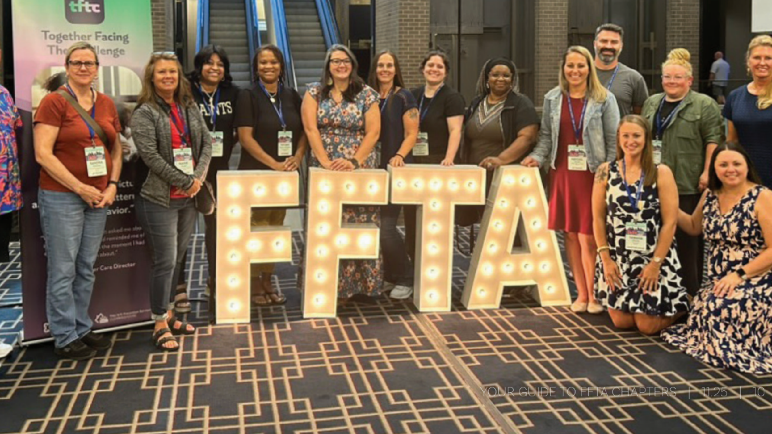 A group of people apart of family services training stand indoors smiling behind large lit-up letters spelling "FFTA."