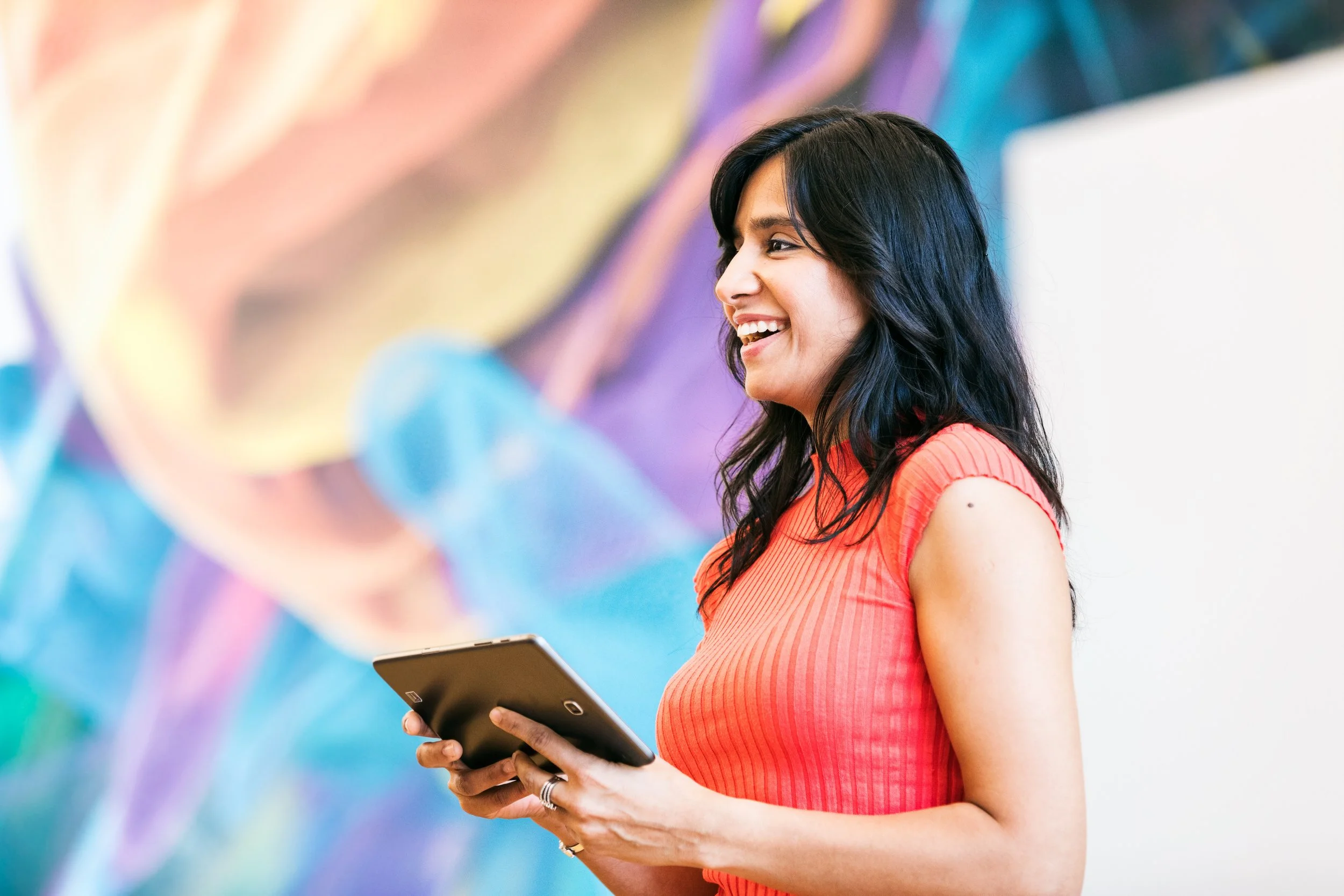 A smiling woman in a red sleeveless top holding a tablet, standing in front of a colorful abstract mural.