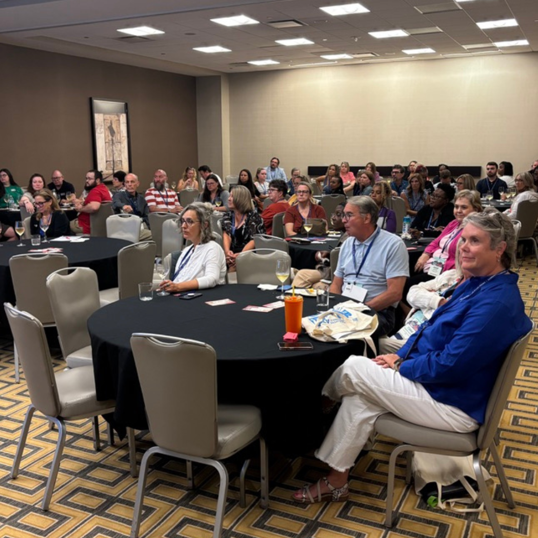 A group of adults seated at round tables attentively listening to a presentation about child and family services in a conference room.