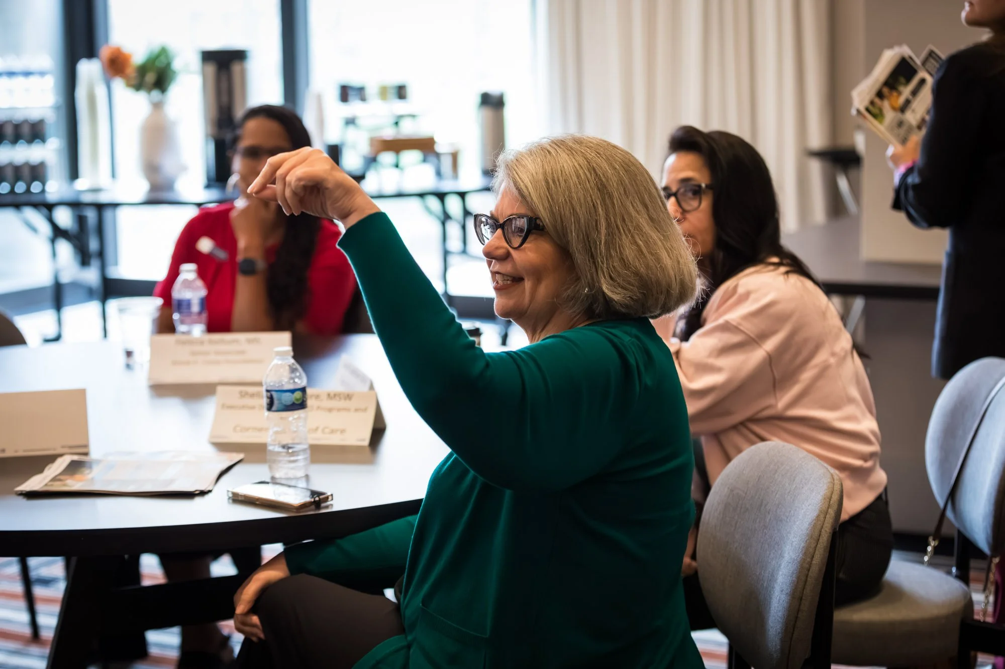 A woman in a green jacket raises her hand while seated at a round table during a meeting about Nonprofit leadership development.