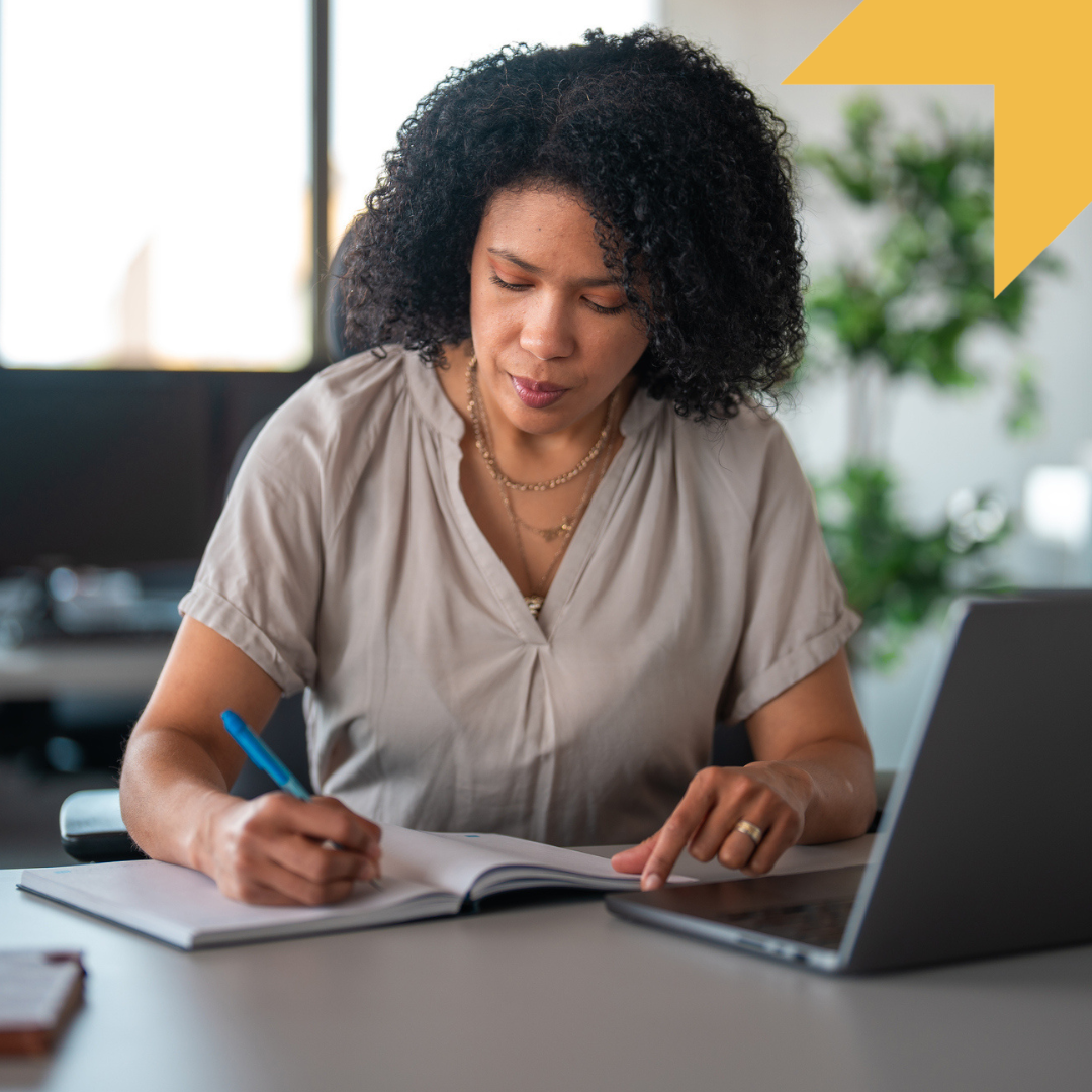 Woman with curly hair writing in a notebook about child welfare leadership at a desk with a laptop, in a bright office setting.