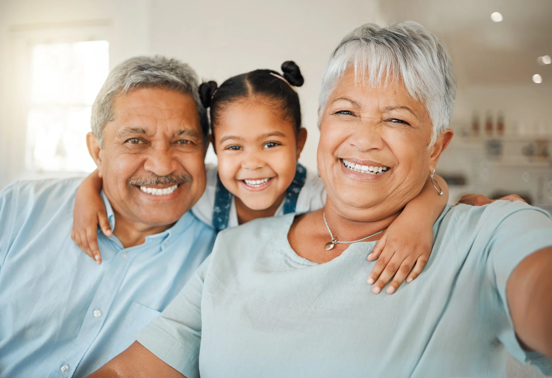Three family members, an elderly man and woman, and a young girl, smiling and hugging each other indoors.