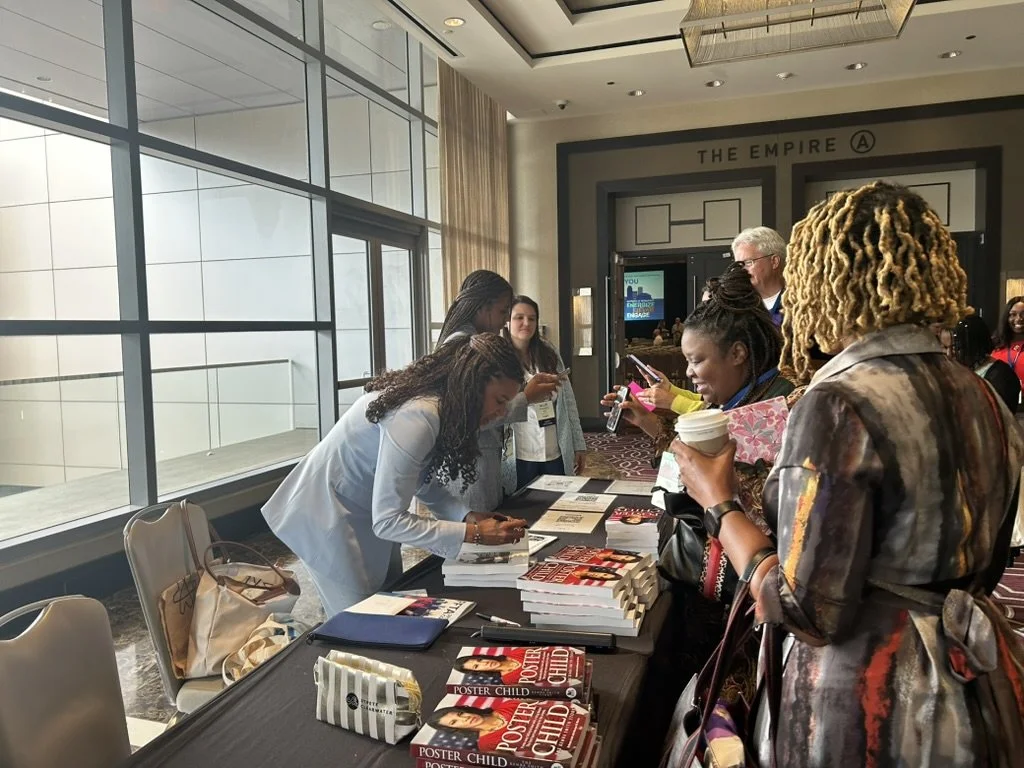 Several people stand at a table with books, while one woman signs a book for another in a conference setting.