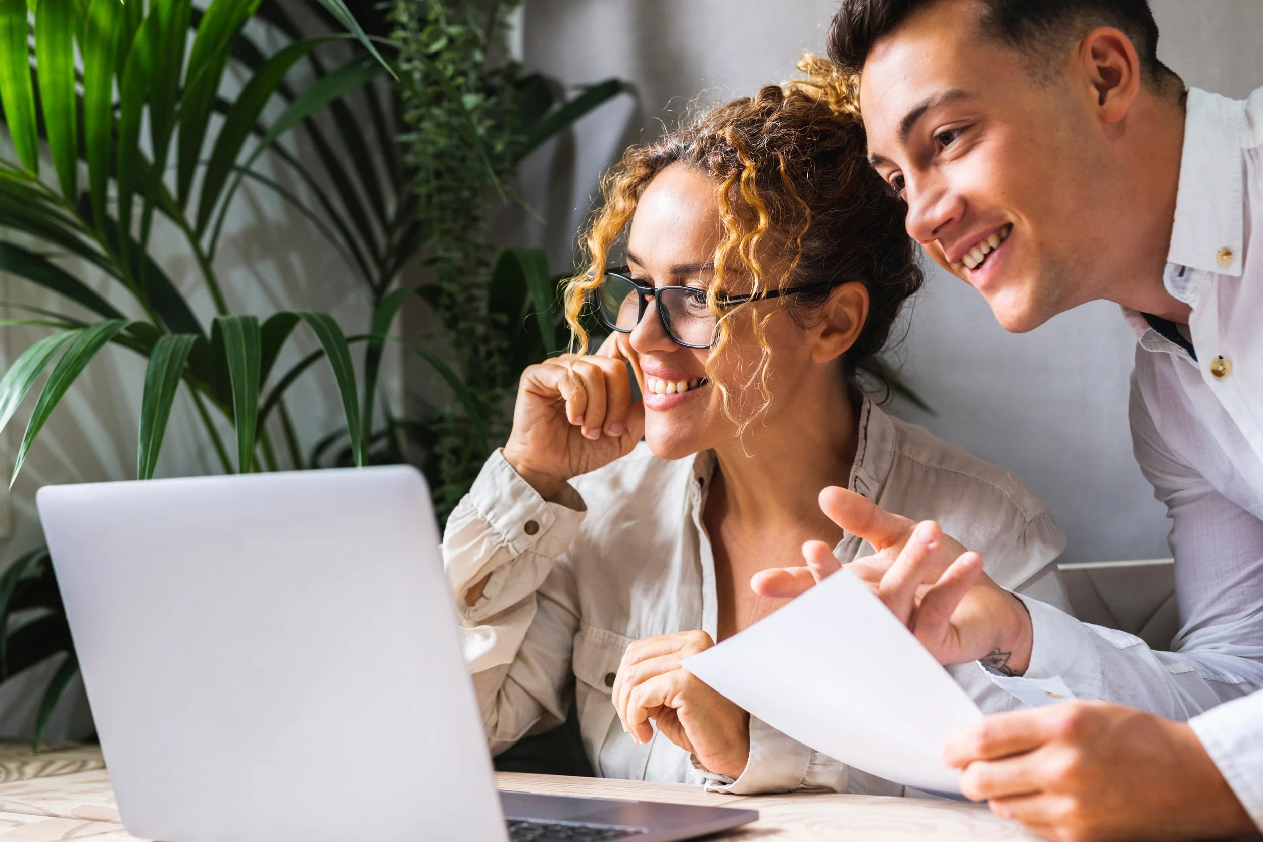 Two people sit at a table, smiling and looking at a laptop screen showcasing child welfare professional development, with papers in hand and plants in the background