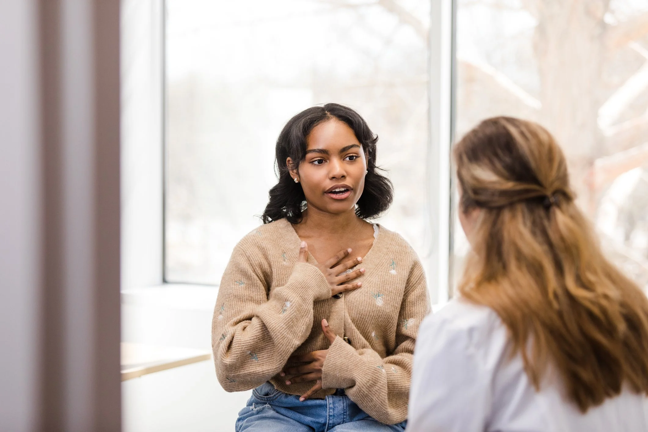 A woman with dark hair, wearing a beige sweater with small embroidered birds, appears to be speaking to another woman with light brown hair in a ponytail. They are indoors next to a large window with a blurred outdoor view.