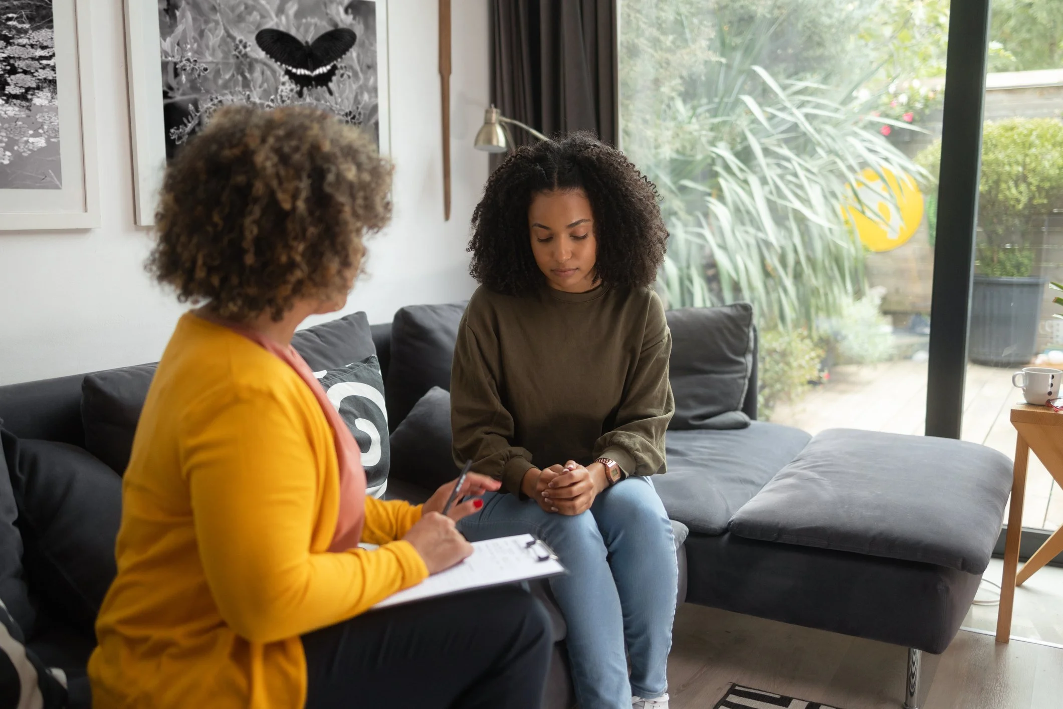 Two women sitting on a black sofa in a living room, engaged in conversation. One woman with curly hair wearing a yellow jacket and holding a notebook, the other with dark hair and wearing a green top. Behind them is a large window with greenery outside, and a black and white butterfly artwork on the wall.