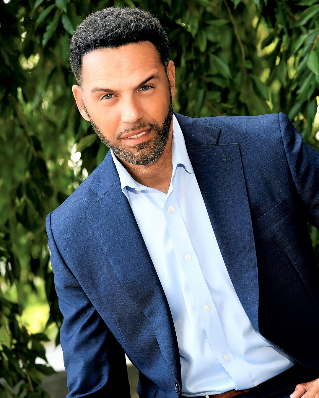 Steve Pemberton in a blue suit jacket and light blue shirt standing outdoors in front of green leafy background.