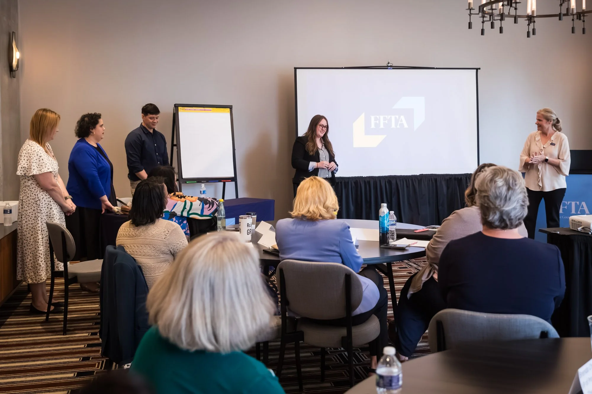 A group presents at the front of a conference room to seated attendees, with an FFTA slide displayed on a screen.