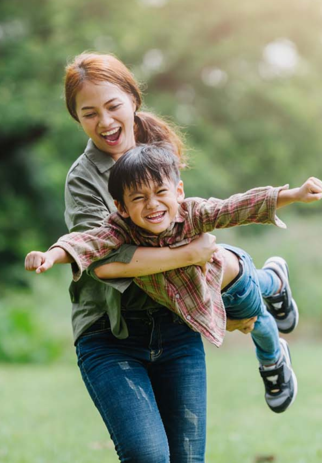 Woman lifting and spinning a smiling young boy outdoors on a grassy field, both appearing joyful representing child and family services.
