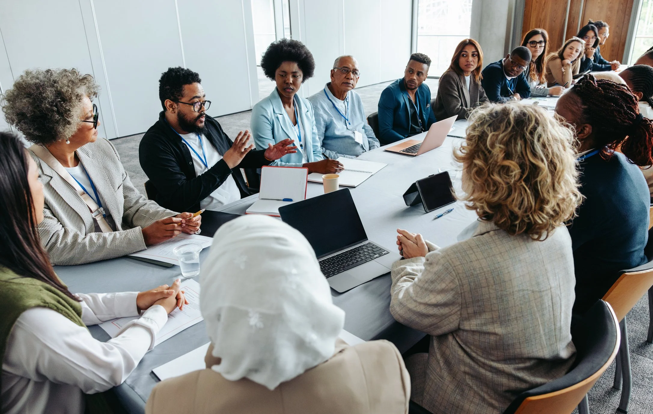 A diverse group of people in business attire attending a meeting in a modern conference room with large windows. Some are taking notes, while others listen attentively or speak.