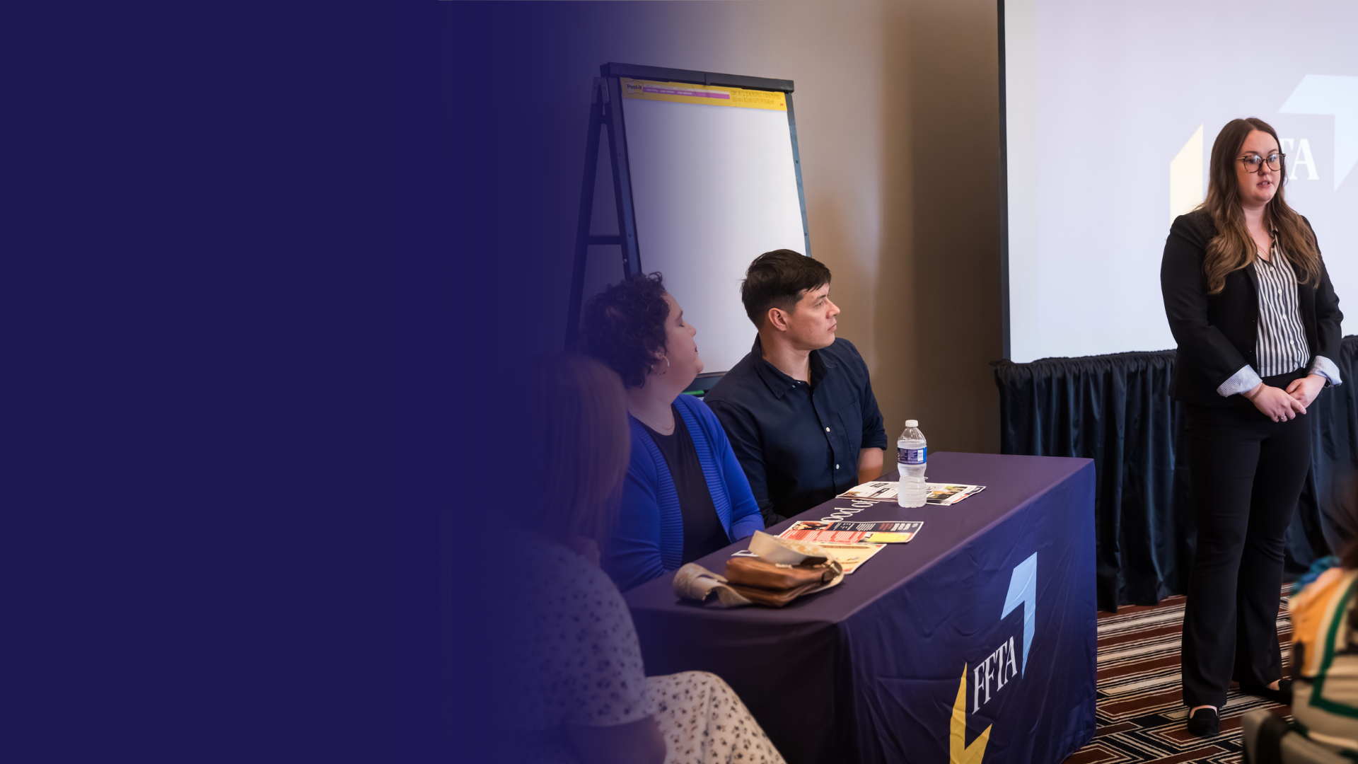 A woman speaks at a conference while three people sit at a table with FFTA branding, facing the audience.