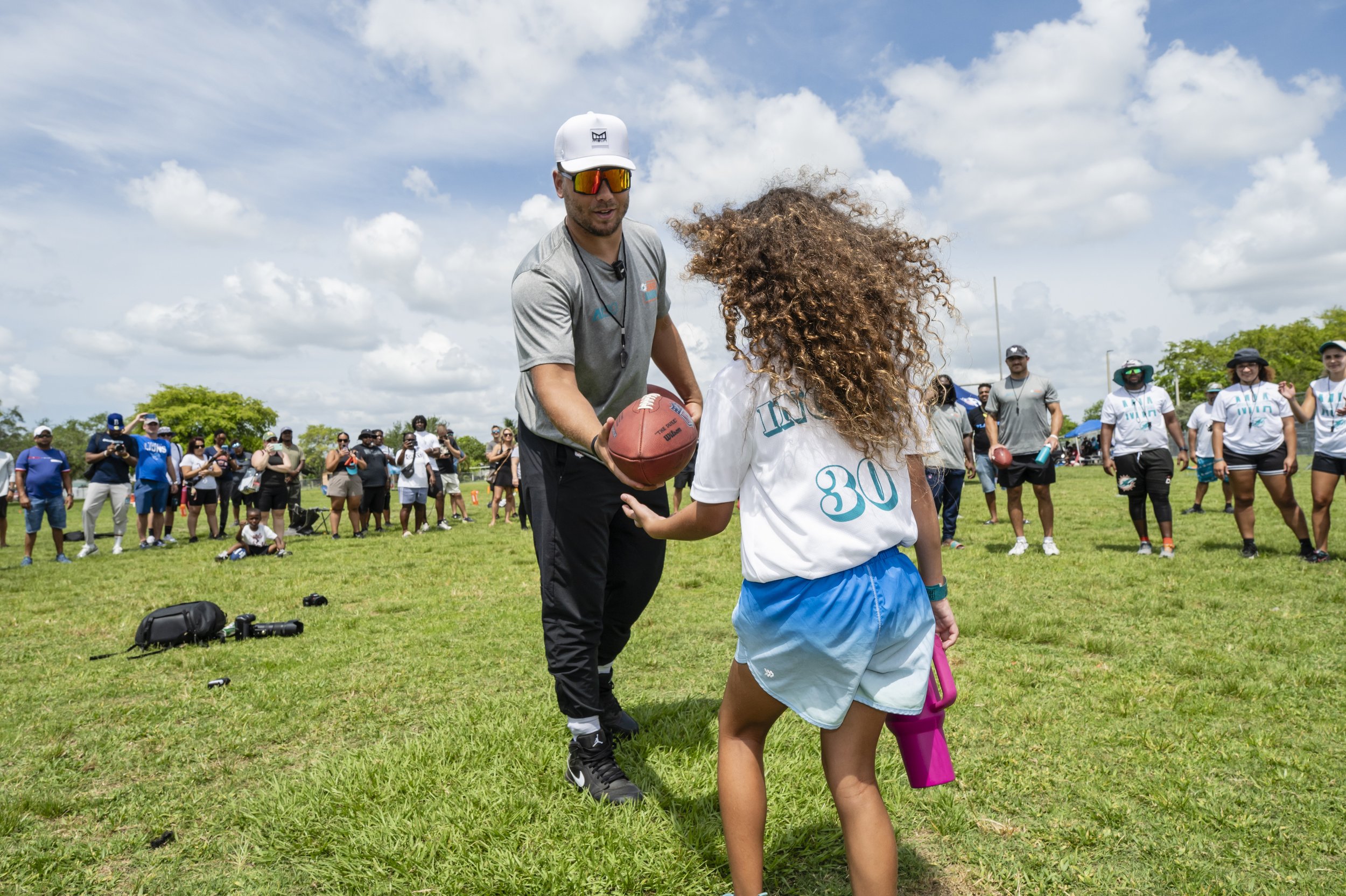 Alec Ingold, a FFTA guest speaker, hands a football to a young girl on a grassy field as a crowd watches in the background.