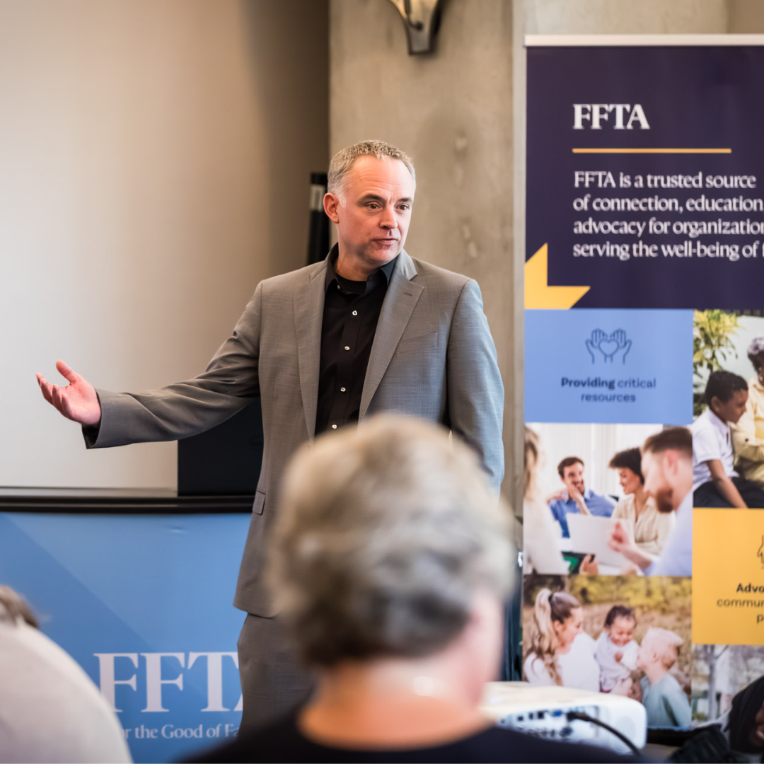 A man in a suit speaks to an audience near an FFTA banner at an indoor event or conference.