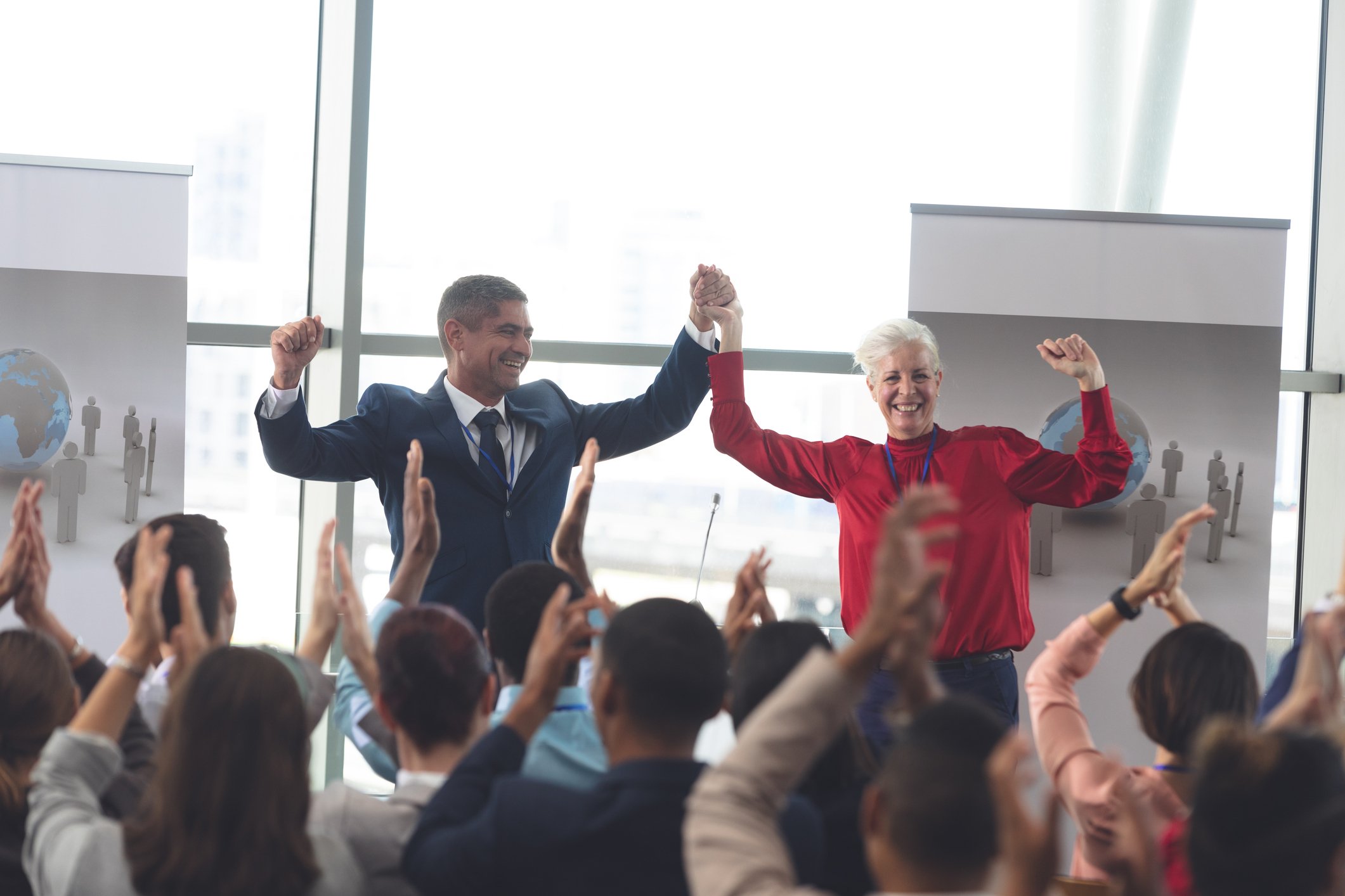 Two businesspeople talking about Kinship care resources, stand holding hands raised in front of an applauding audience at a conference or seminar.