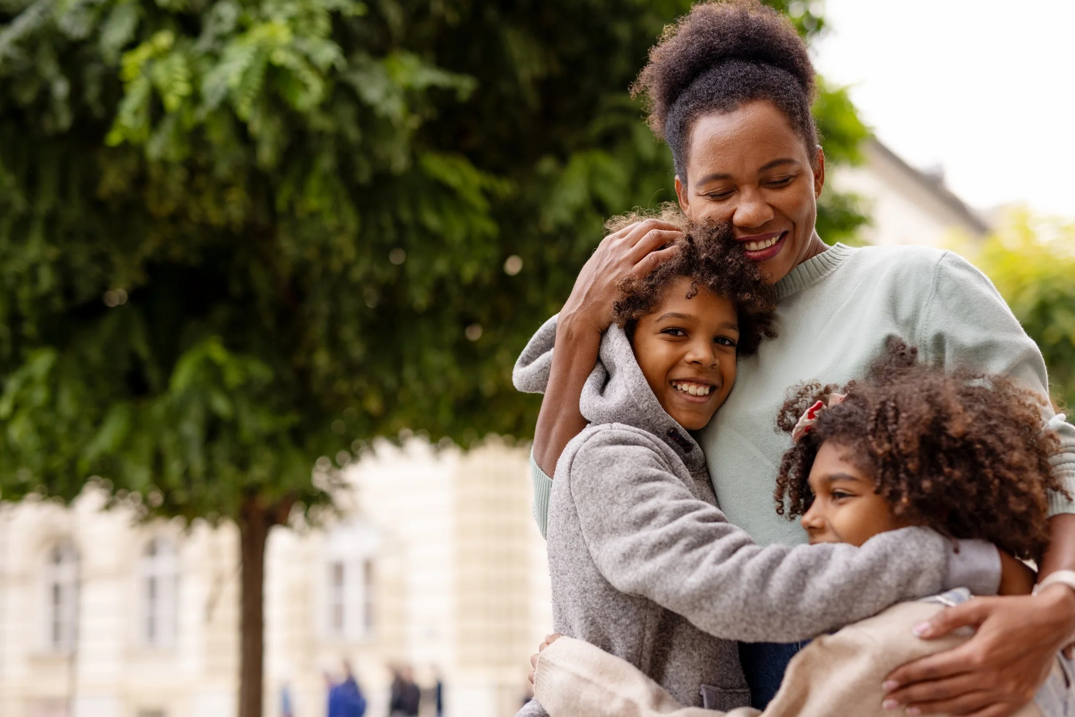 A woman hugging a young girl with curly hair outside with a blurred background of trees.