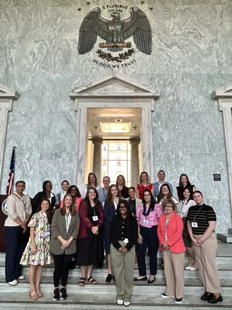 A group of child welfare professionals poses for a photo on marble steps beneath an eagle emblem and flags in a grand hall.