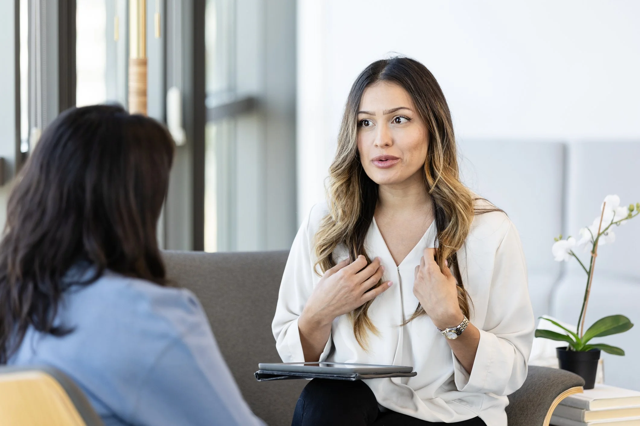 A woman with long, wavy hair and a white blazer is speaking to another woman with dark hair, in a professional setting with a white wall and a potted plant in the background.