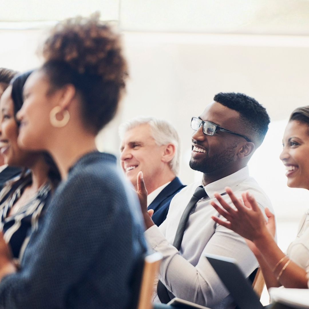 A group of people in business attire sit and applaud at an indoor presentation discussing family well-being organizations.