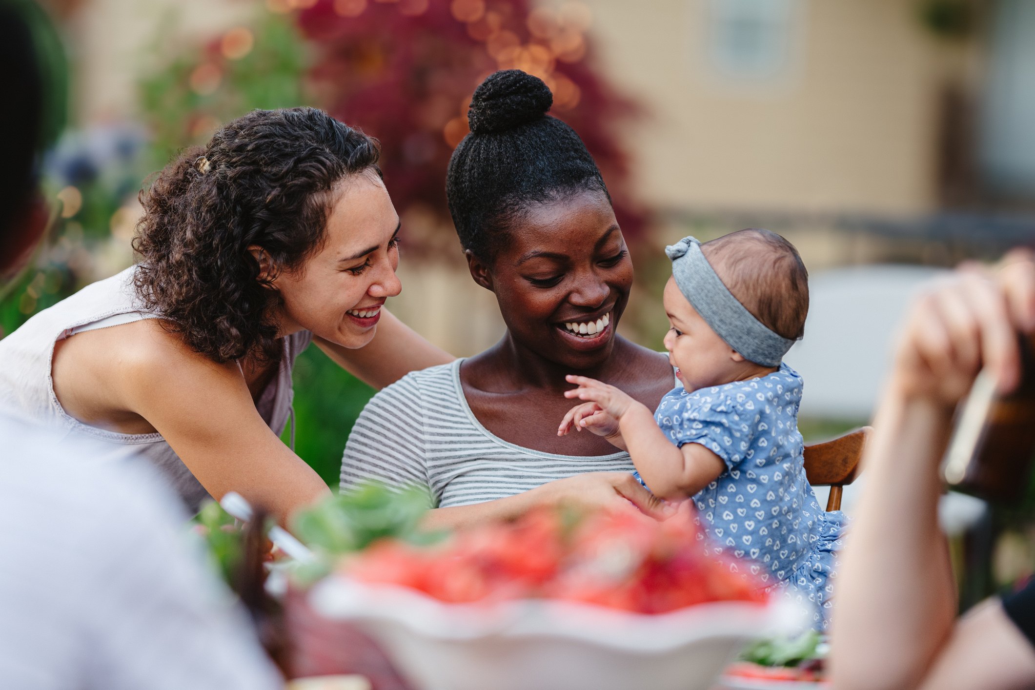 Three women and a baby are smiling and interacting outdoors, possibly at a social gathering or family event, with colorful plants and blurred background.