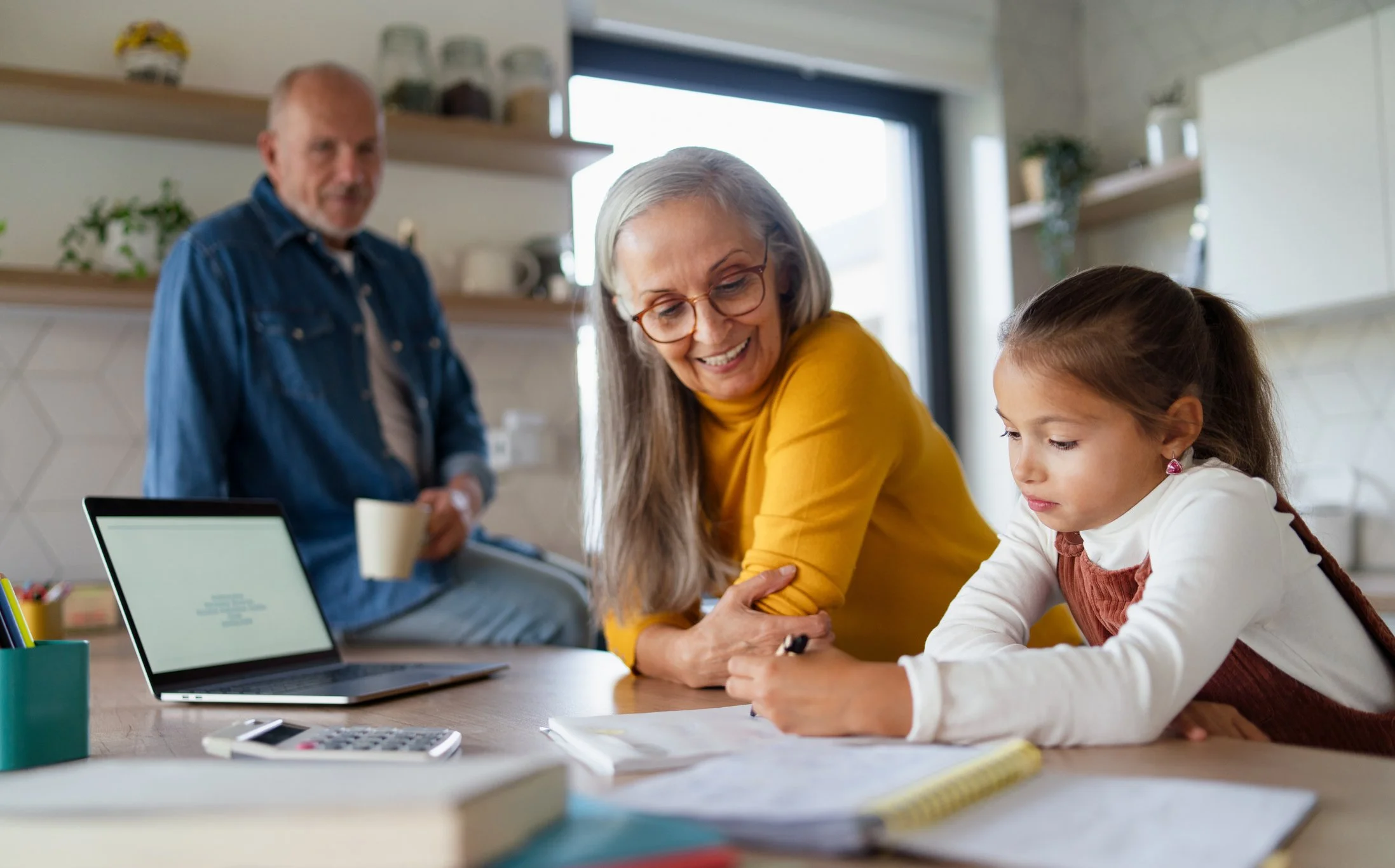 An elderly woman in glasses and a yellow shirt is helping a young girl with homework at a table, with a man in a blue shirt standing in the background in a kitchen.