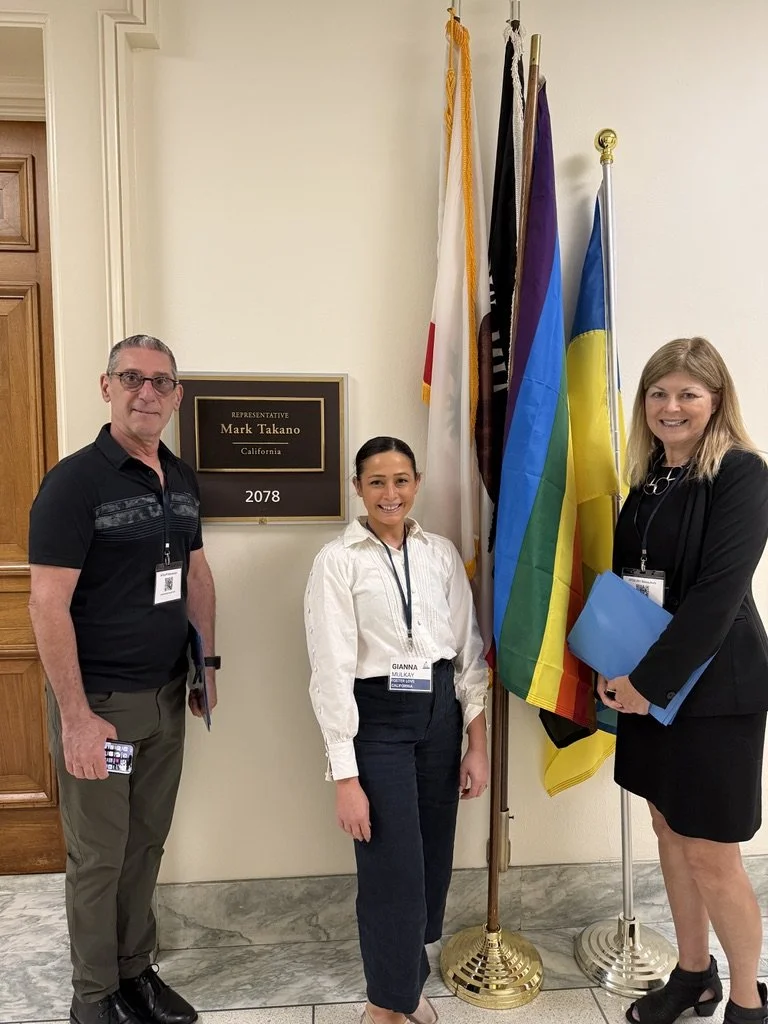 Three people stand by flags and a sign for Representative Mark Takano outside office 2078 in a hallway.
