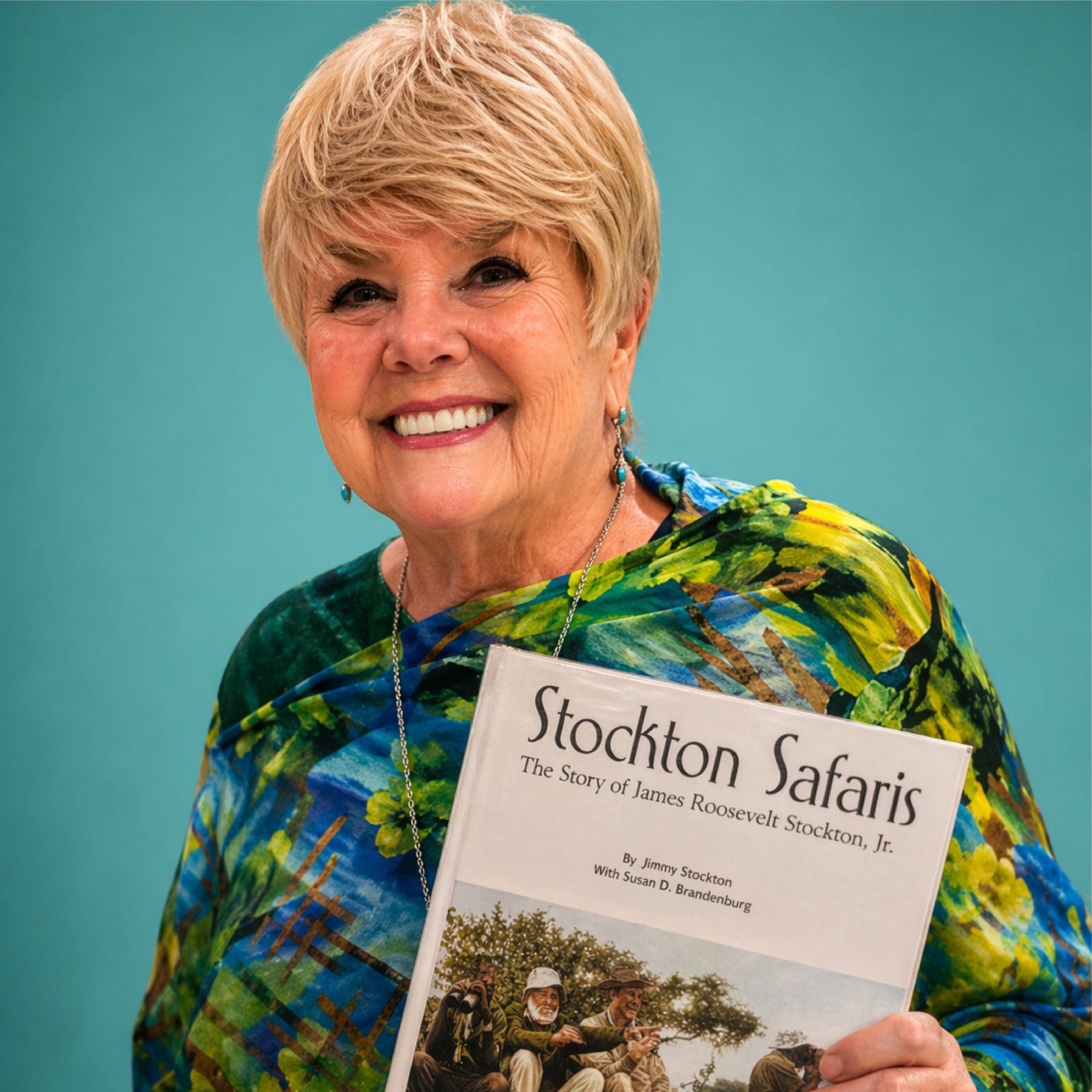 Close-up of Susan Brandenburg with short blonde hair smiling, wearing colorful clothing and holding a book titled 'Stockton Safaris: The Story of James R. Roosevelt Stockton, Jr.' against a teal background.