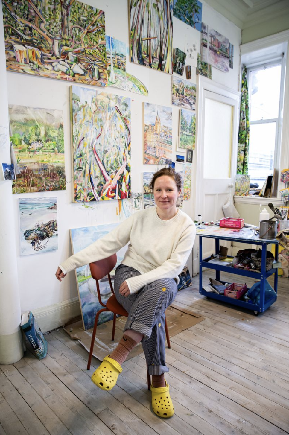 A woman in a white sweater and striped pants wearing yellow Crocs sitting in an art studio surrounded by colorful paintings on the wall and sitting on a red chair. There is a blue cart with art supplies nearby and natural light coming from a large window.