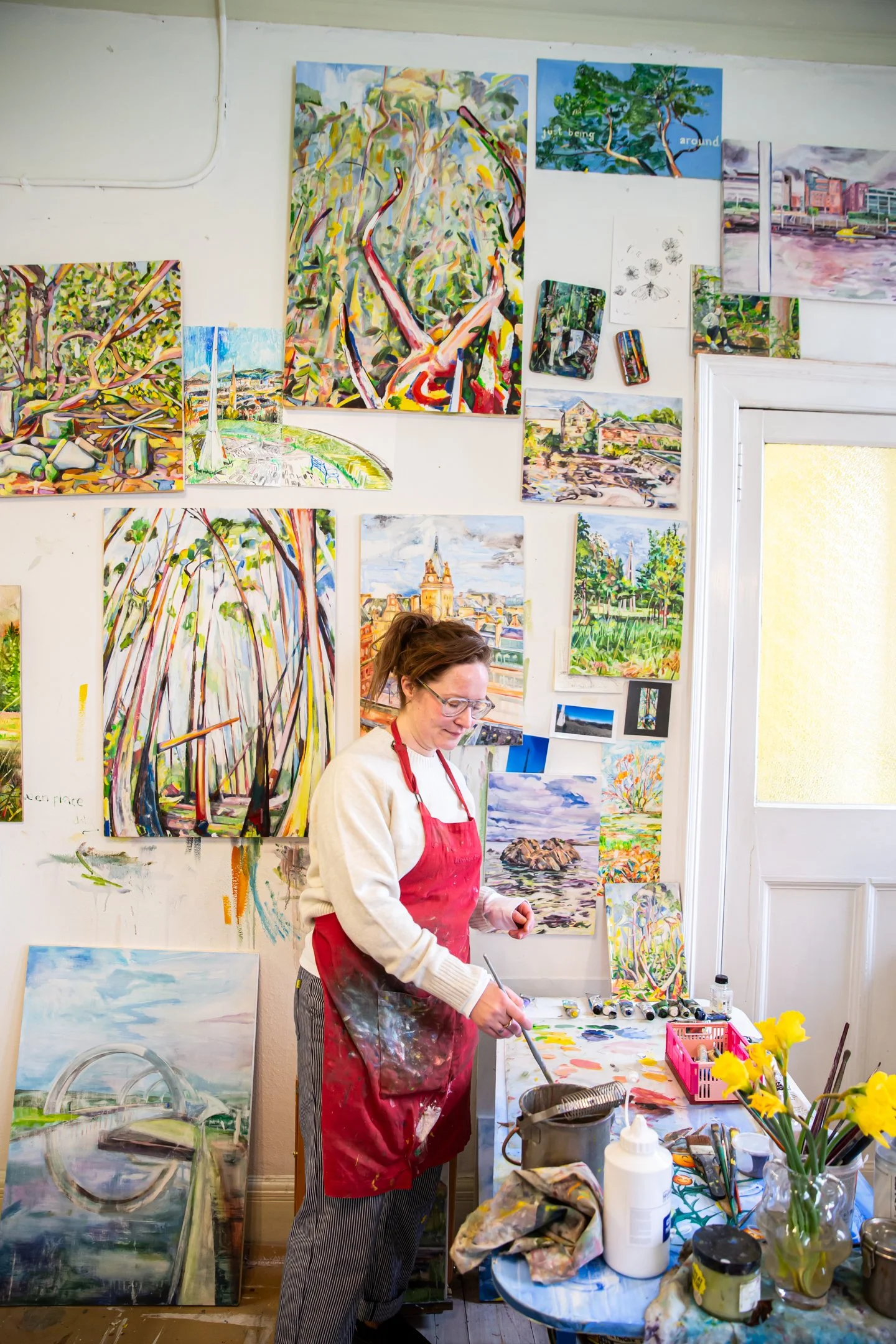 An artist painting in her studio. The wall behind her is decorated with various colorful landscape and abstract paintings. The table in front of her has paint supplies, brushes, and flowers.