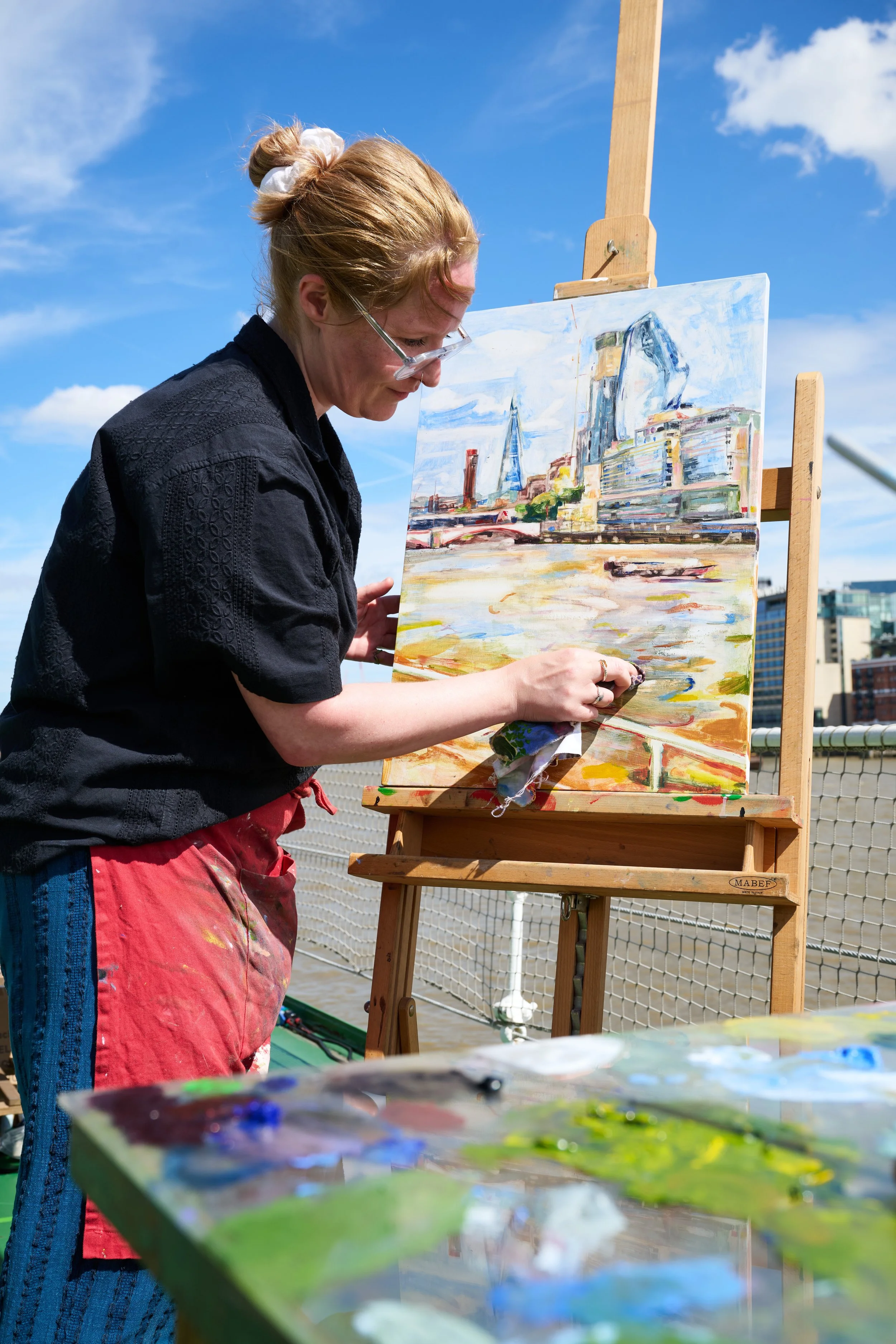 A female artist painting a cityscape during the daytime on an outdoor rooftop, with a bright blue sky and some clouds in the background.