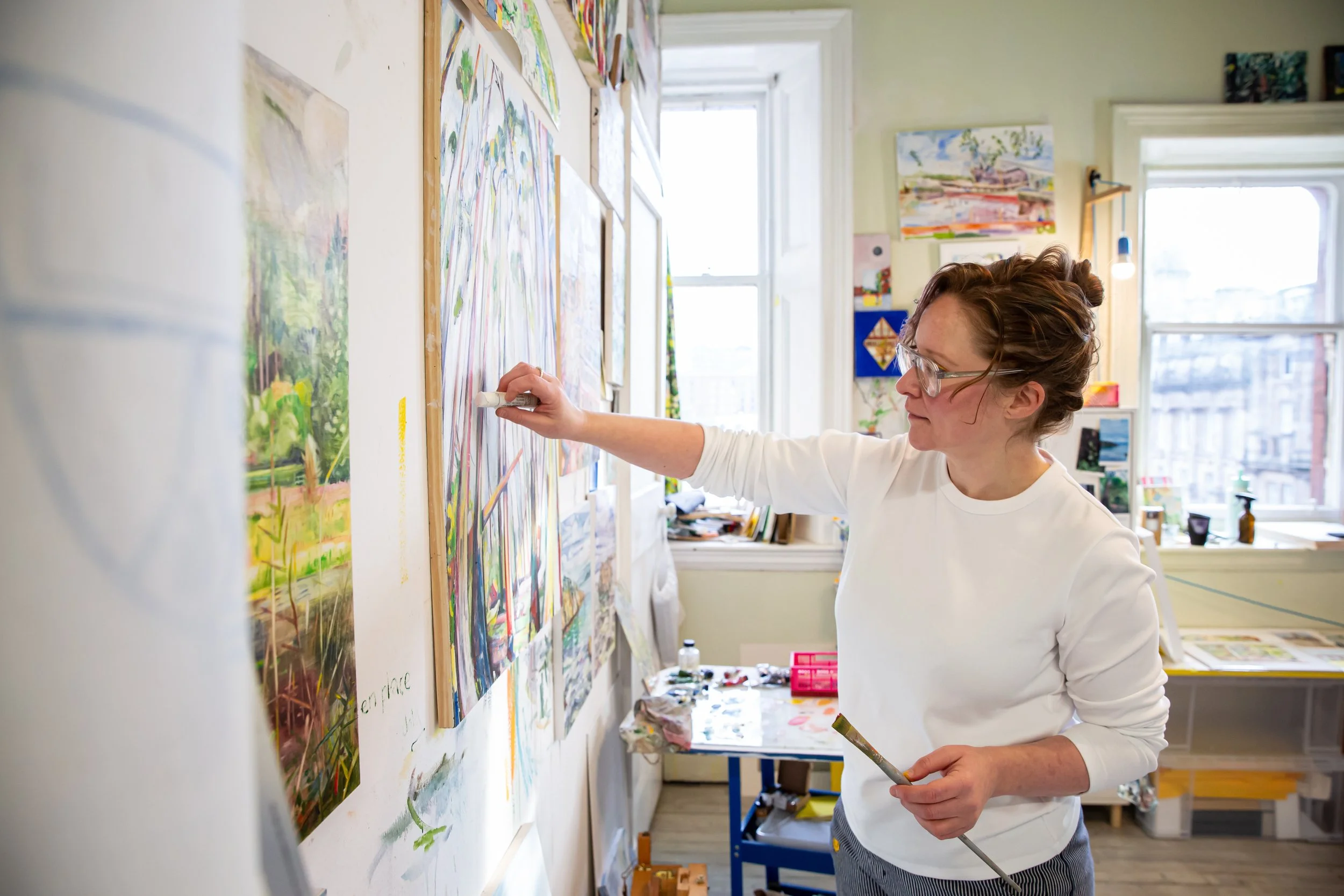 A woman wearing glasses and a white long-sleeve shirt painting colorful artwork on a wall in an art studio, with windows and various paintings and supplies visible in the background.