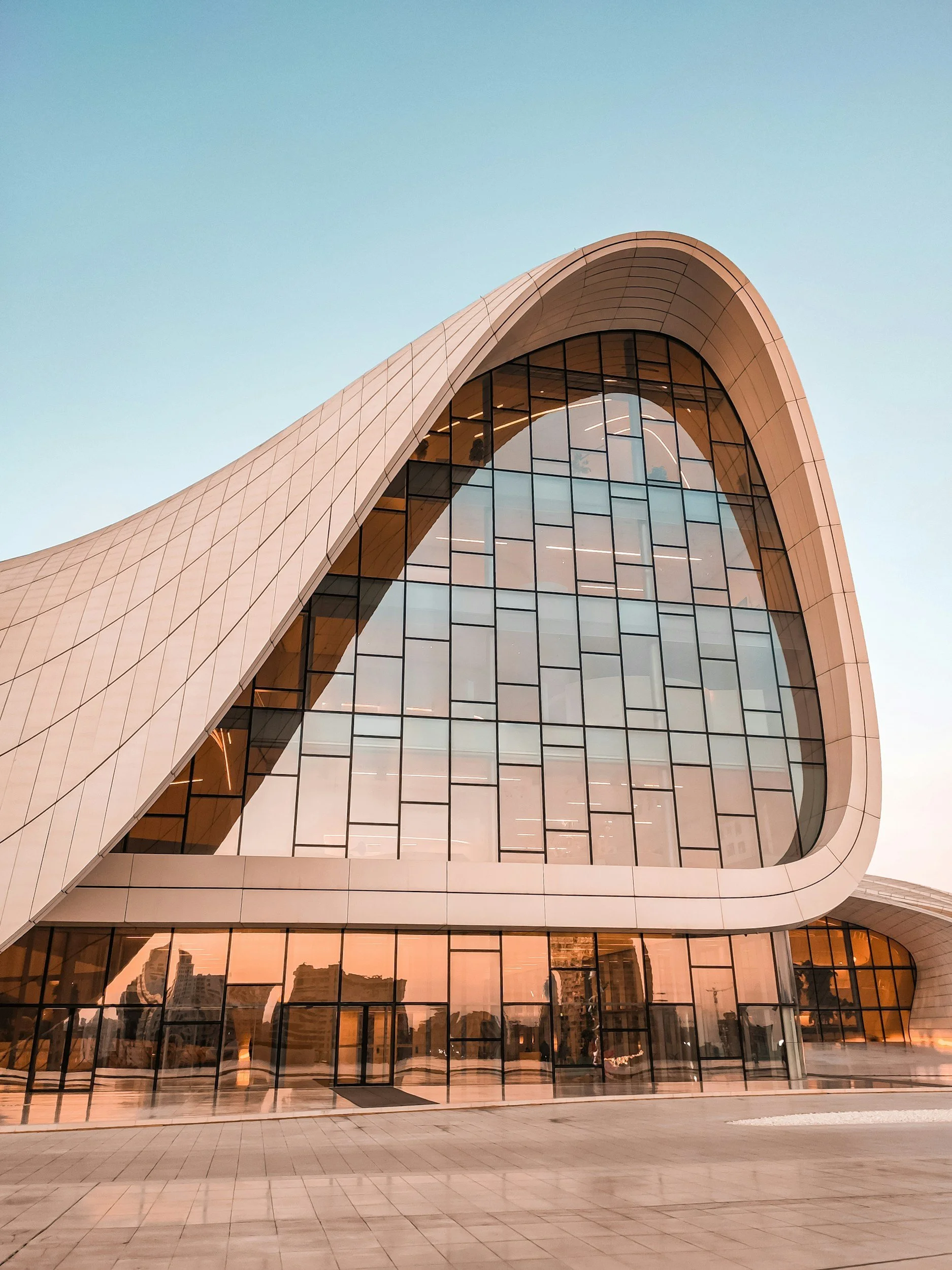 Modern building with a curved, arching roof and large glass windows, reflecting the city skyline and a pink sunset sky.