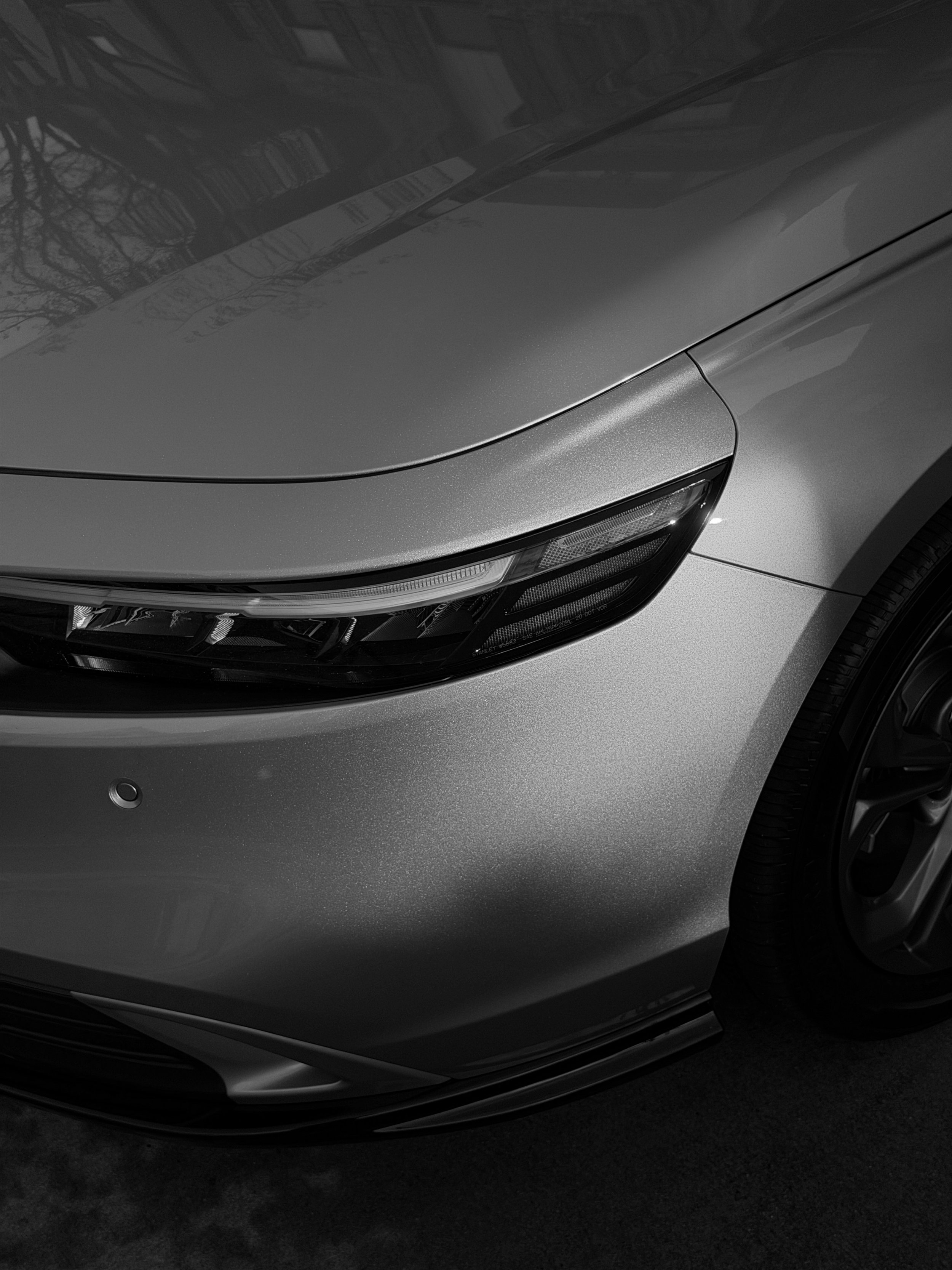 Close-up of the front of a silver car, showing the headlight, bottom bumper, and part of the hood and wheel, with reflections on the surface.