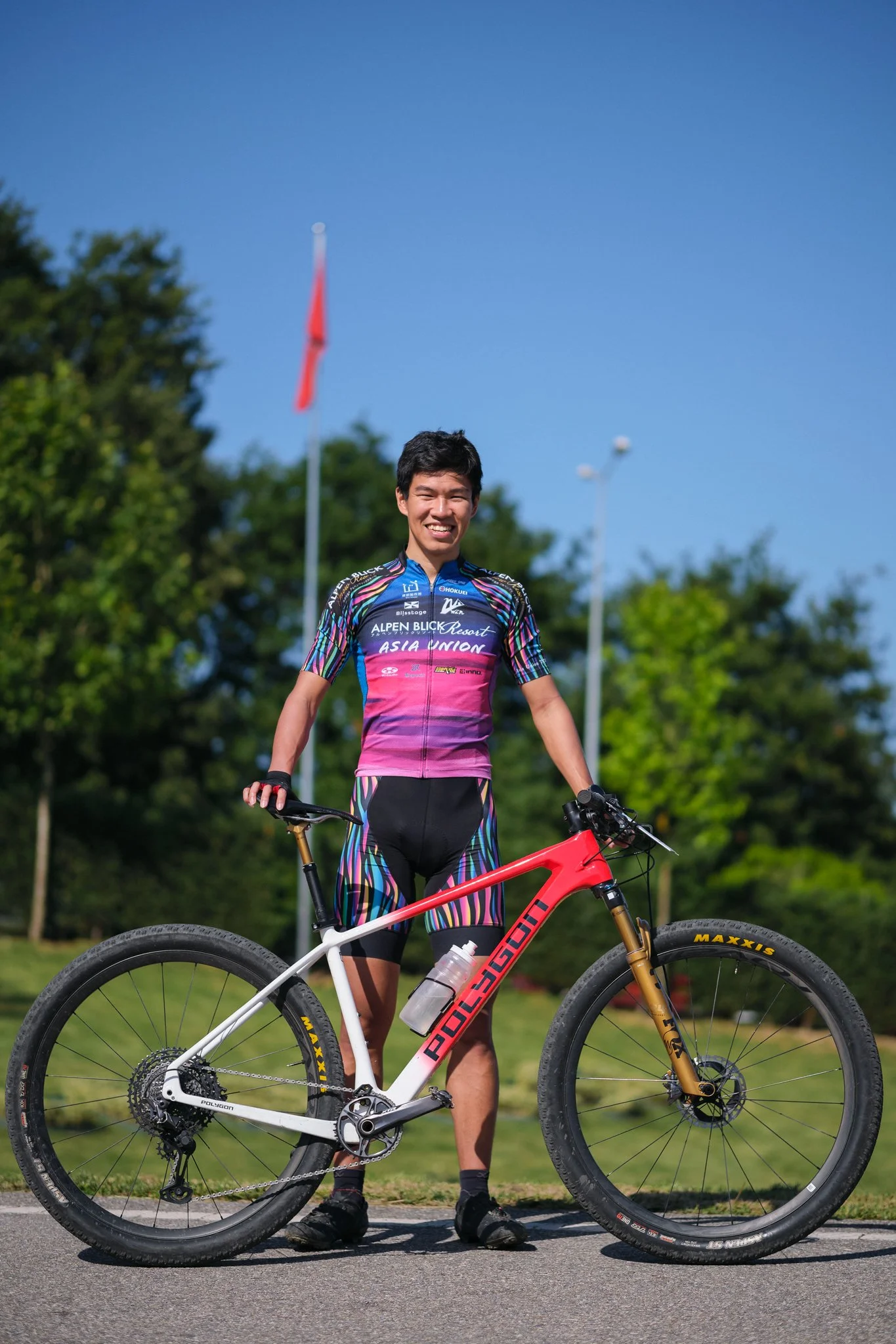A young man in colorful cycling gear standing outdoors with a mountain bike, smiling under a clear blue sky.