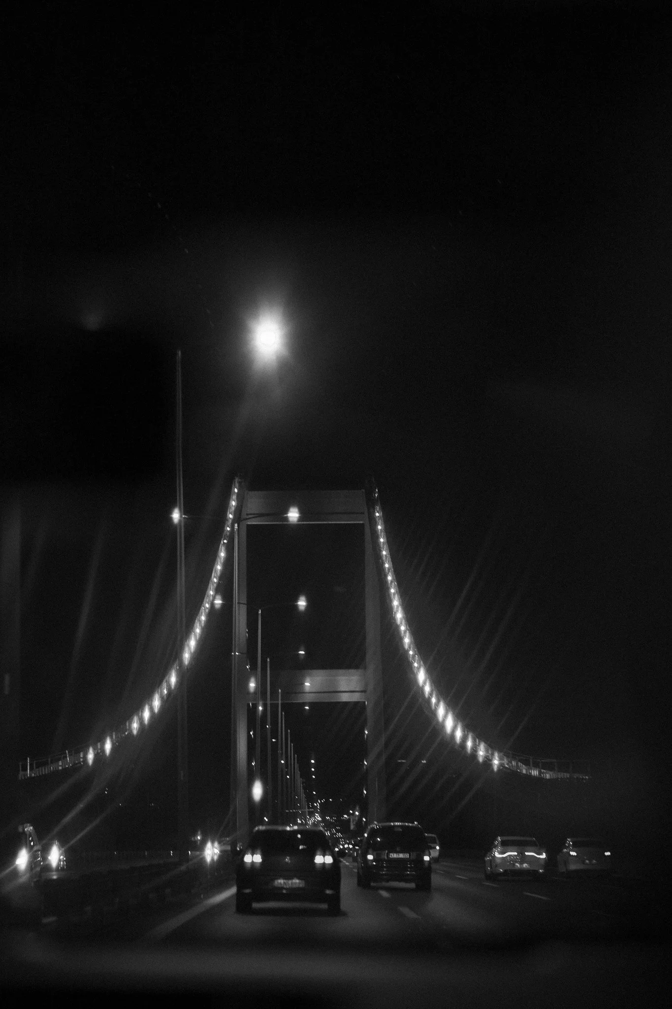 Nighttime scene of a lit-up suspension bridge with cars driving underneath. The bridge's lights and the moon are visible in the dark sky.