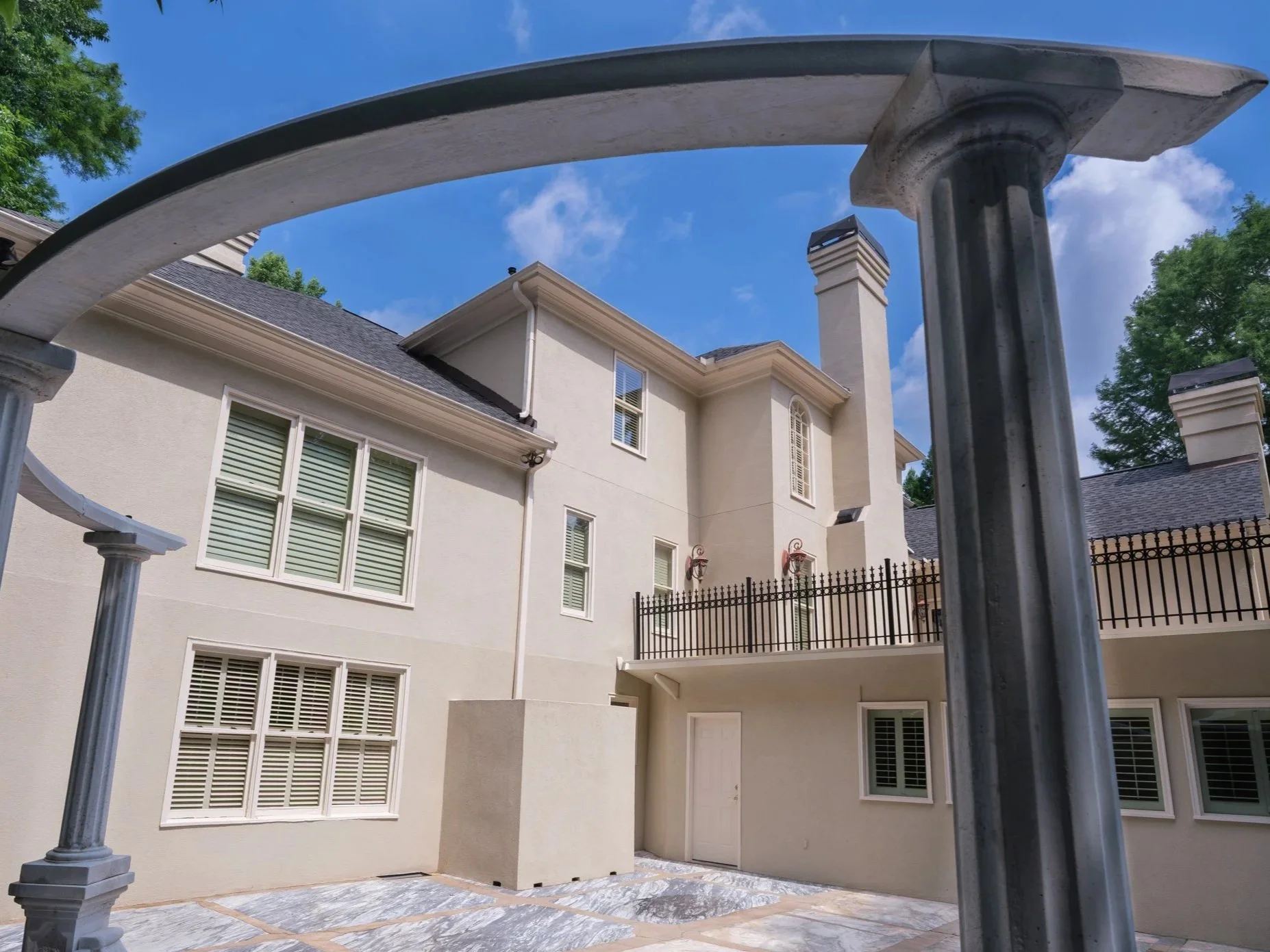 Side view of a large, light-colored residential house with multiple windows, a balcony, and two chimneys. The photograph is taken from under a metal archway in the backyard, with trees visible in the background and a bright blue sky with a few clouds