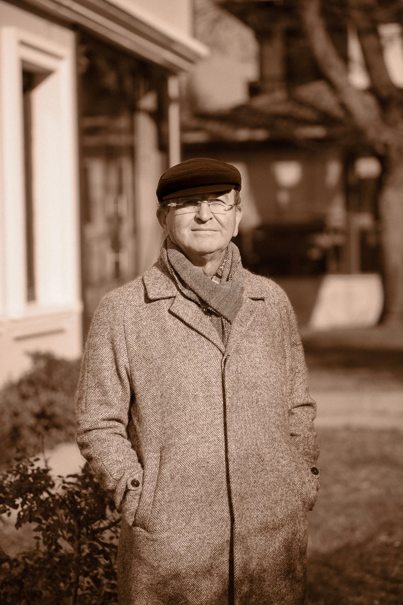 A man in a coat, scarf, glasses, and a hat standing outside in front of a building with trees in the background.