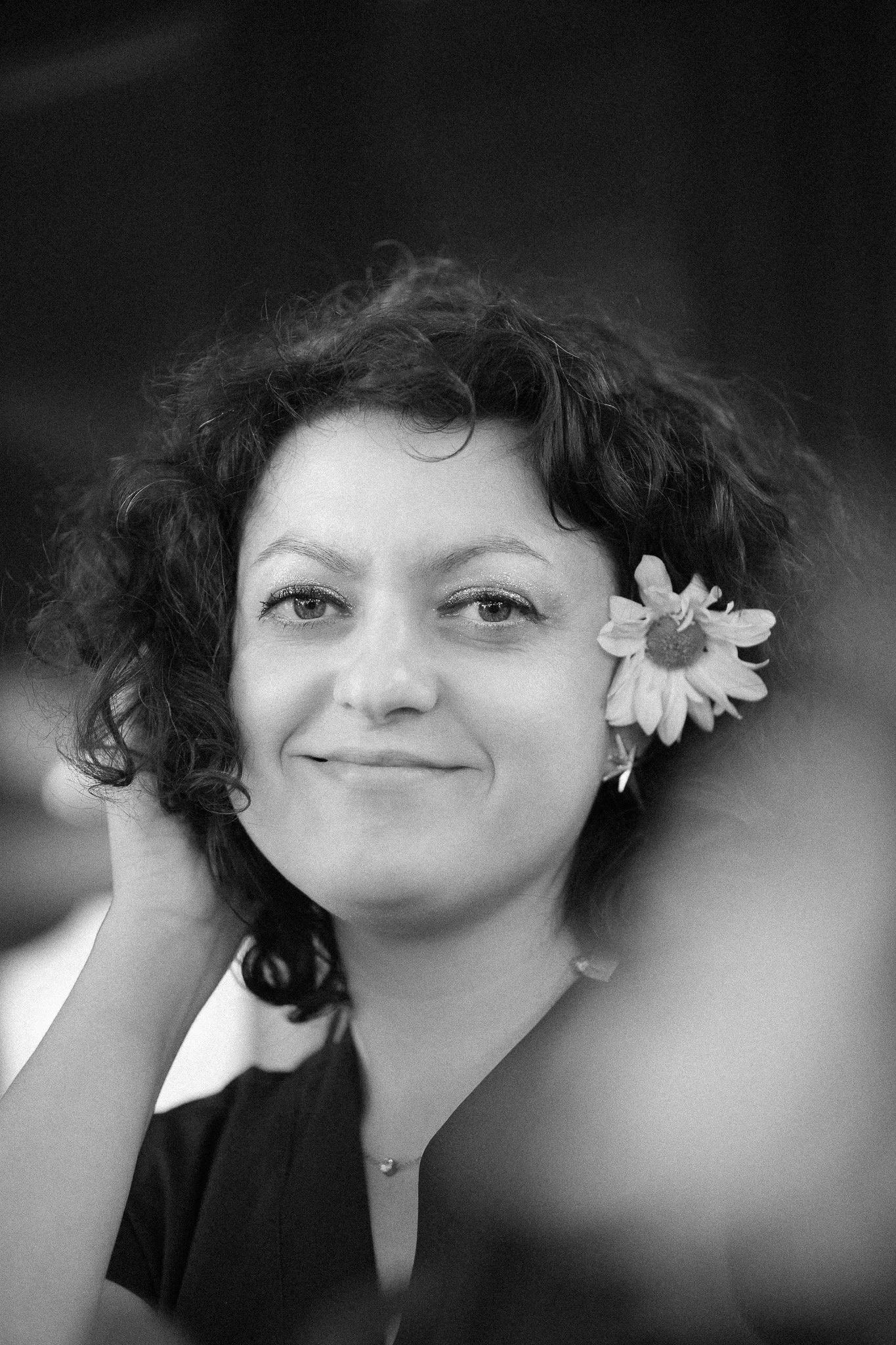 Black and white photo of a woman with curly hair holding a flower near her face, smiling softly.