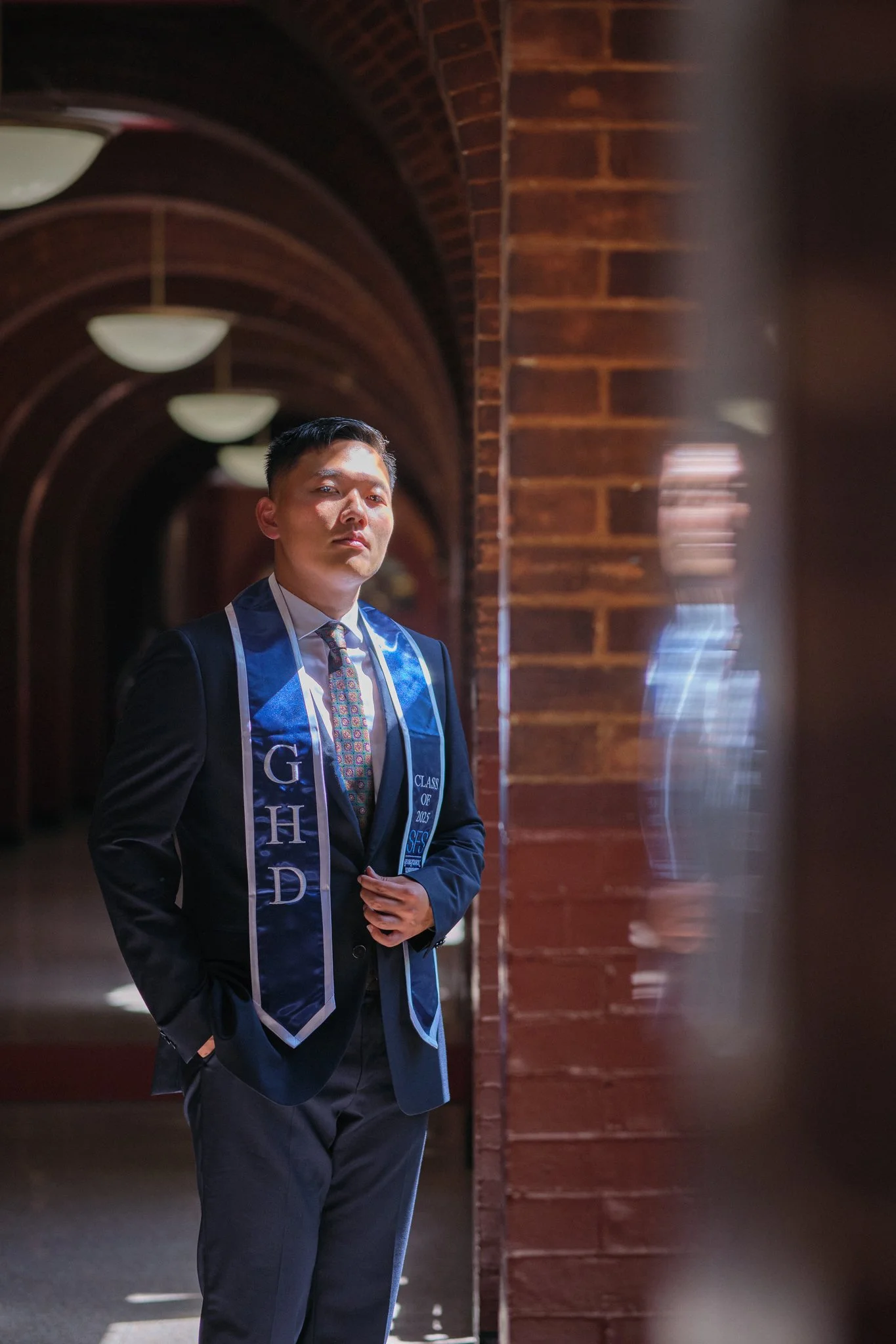 A man dressed in formal attire, wearing a blue graduation stole, standing in a corridor with arched brick walls and hanging lights.