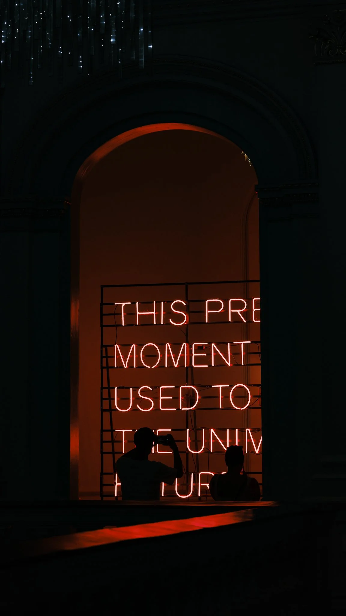 Neon sign displaying the message: 'This pre moment used to tense', with two silhouetted people in front of it, inside a dark building with an arched window and decorative architecture.