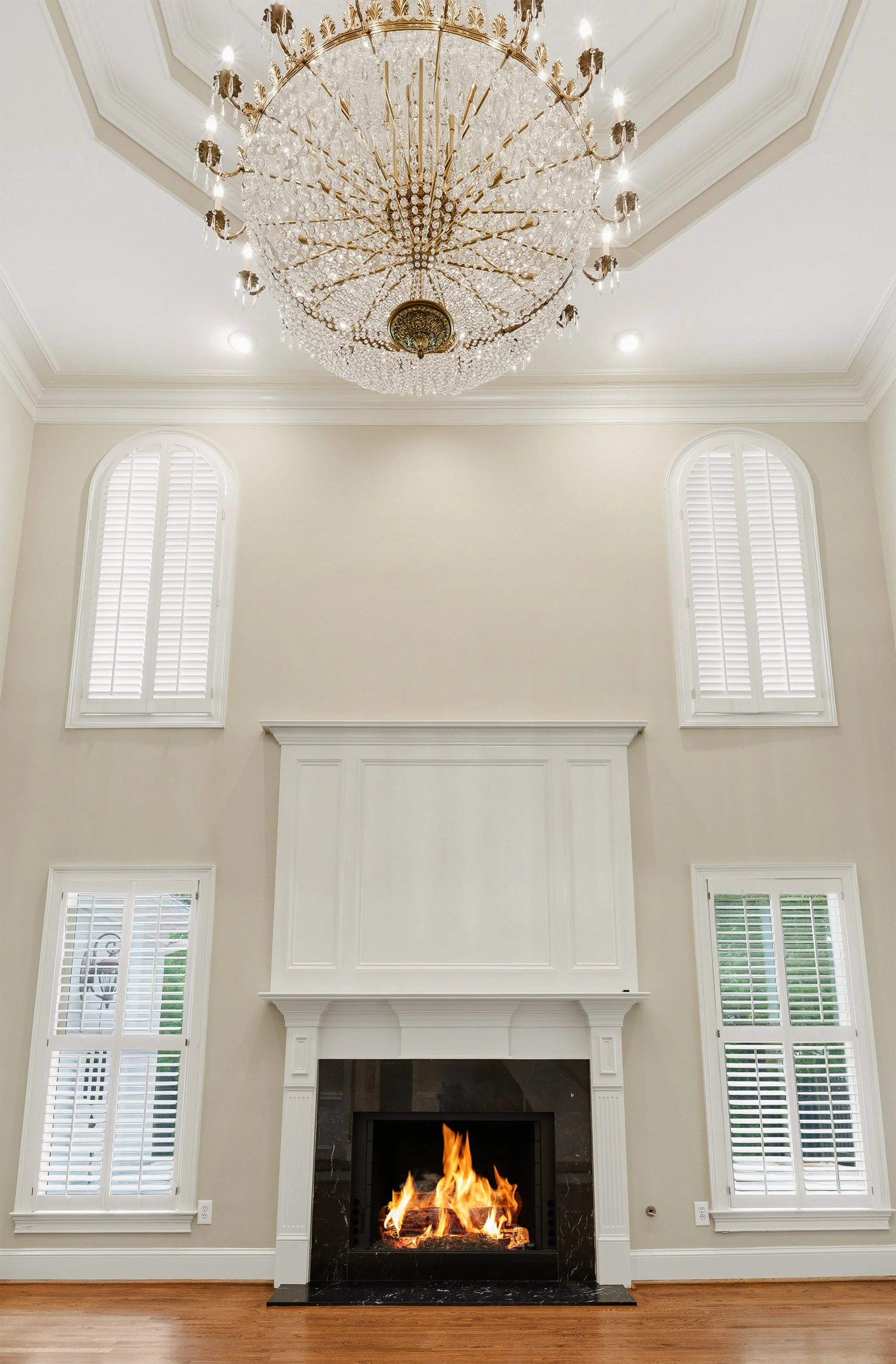 A living room with a white fireplace, two large arched windows with plantation shutters, a large ornate chandelier hanging from the ceiling, and hardwood flooring. Real Estate Photography | Alpharetta | Milton | Buckhead | Atlanta | Drone | FAA Part 